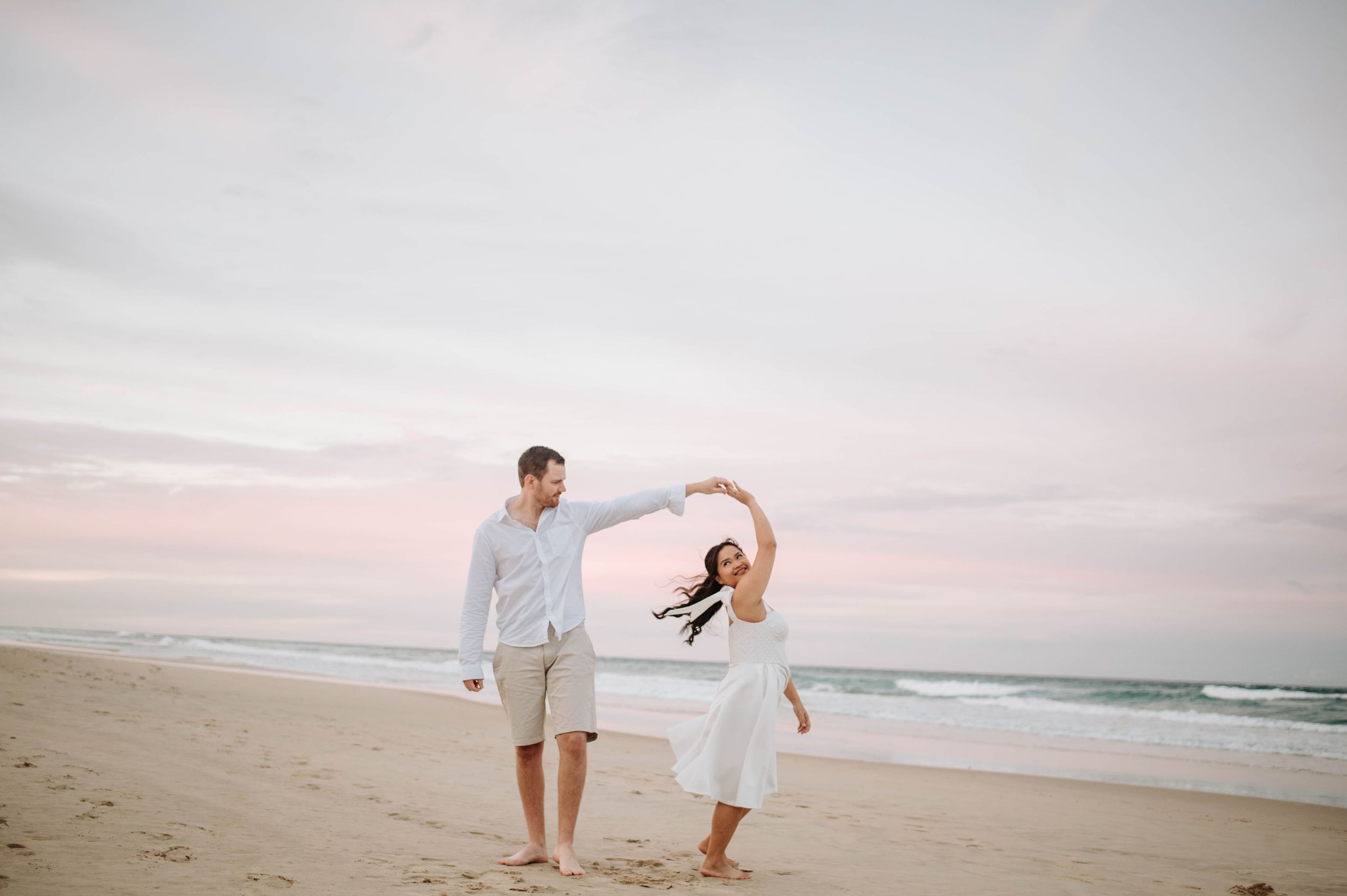 Romantic couple dancing together on beach during sunset with flowing white dress.