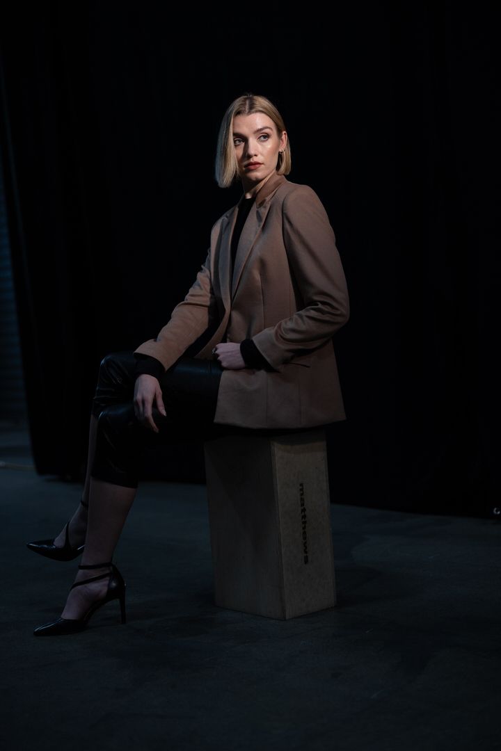 Blonde woman in tan blazer and black leather pants poses on a wooden block against a dark background.