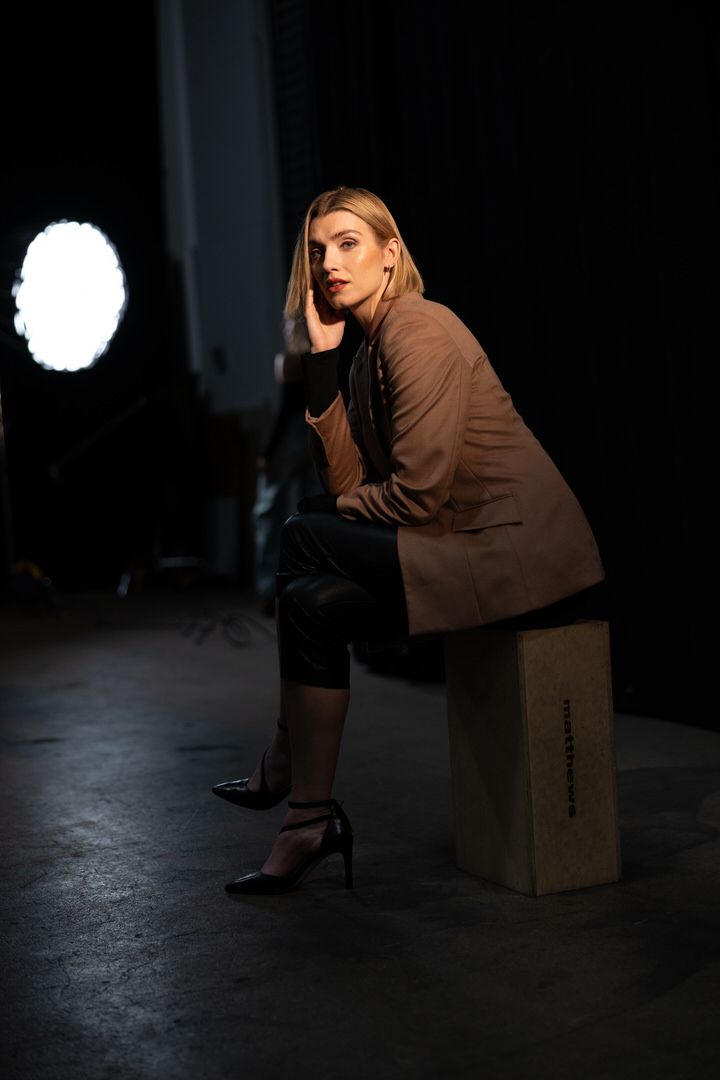 Blonde model in brown blazer sits on a wooden block in a dark studio with a bright light behind her.