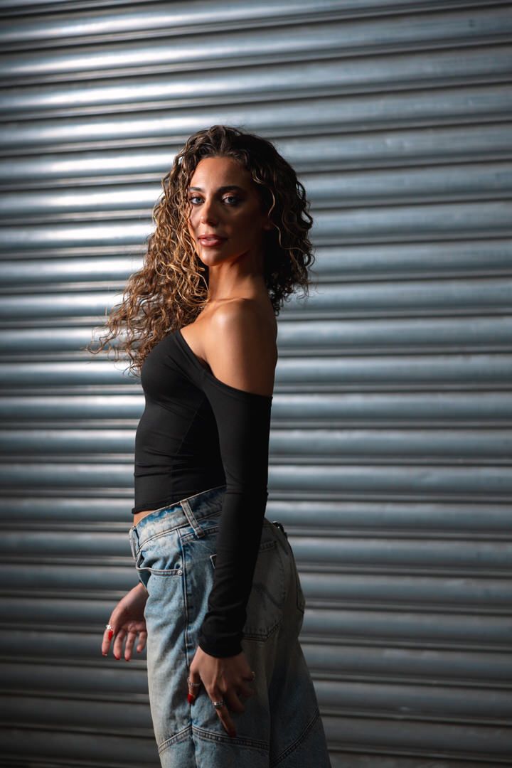 Young woman with curly hair poses in black off-shoulder top and wide-leg jeans against metal shutter.
