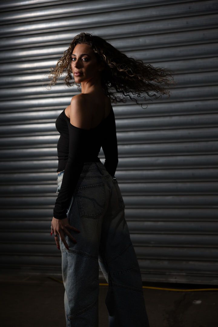 Woman with windswept curly hair poses in black off-shoulder top and light jeans against metal shutter.