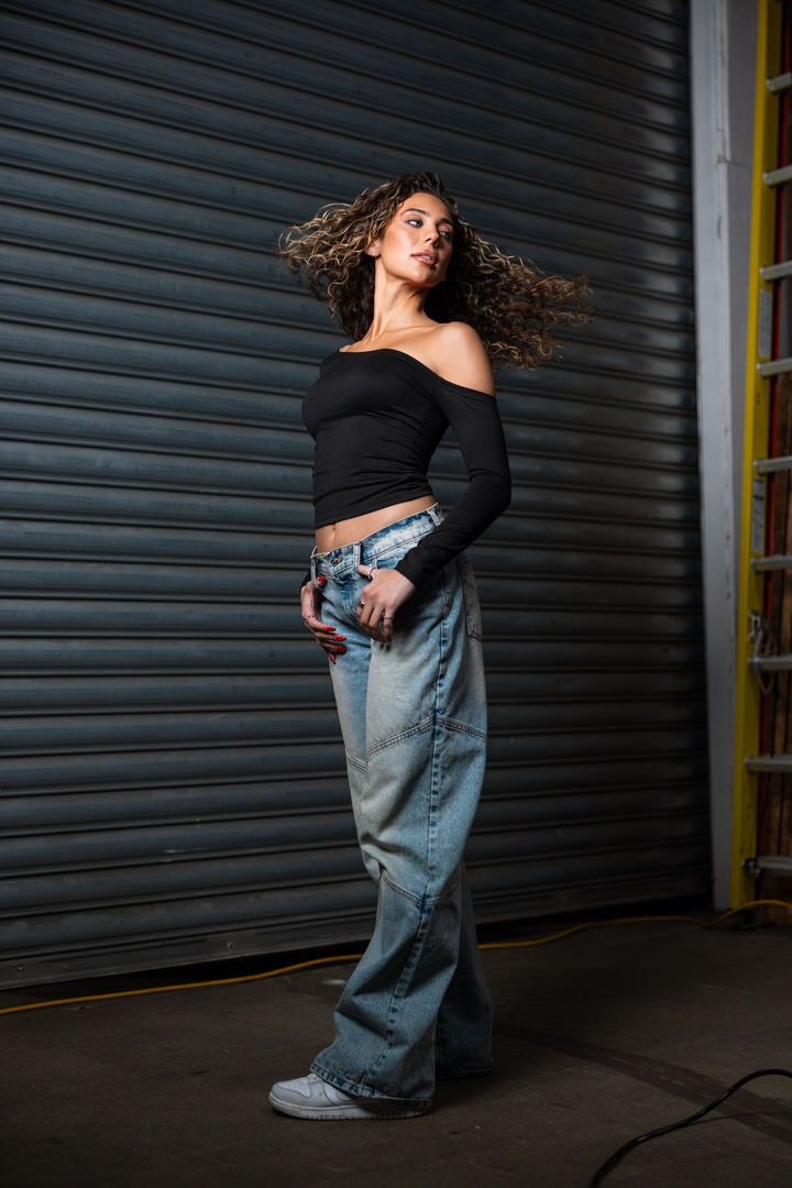 Woman in black off-shoulder crop top and oversized jeans flips hair beside corrugated metal garage door.