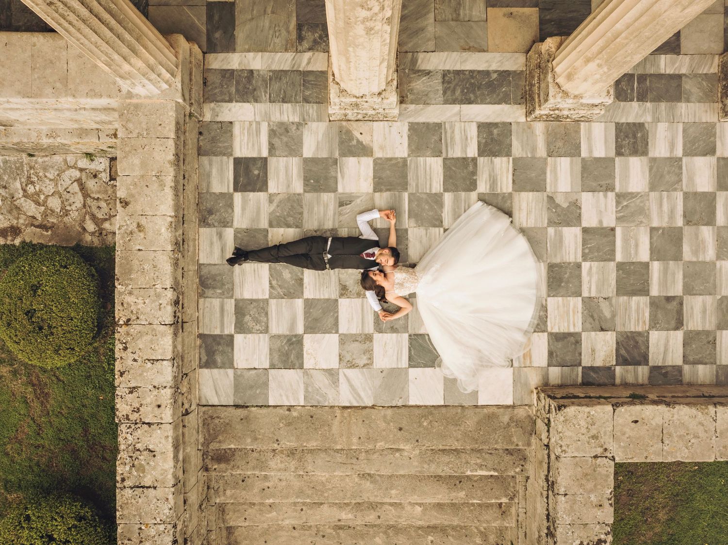 Aerial view of couple in wedding attire standing on checkered tile floor surrounded by columns and grass.