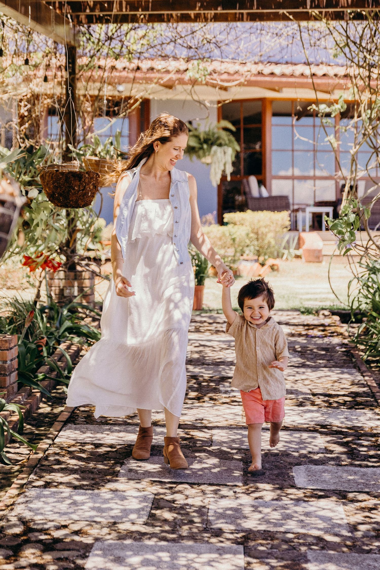 A smiling woman in a white dress walks hand-in-hand with a young child through a sunny garden path.