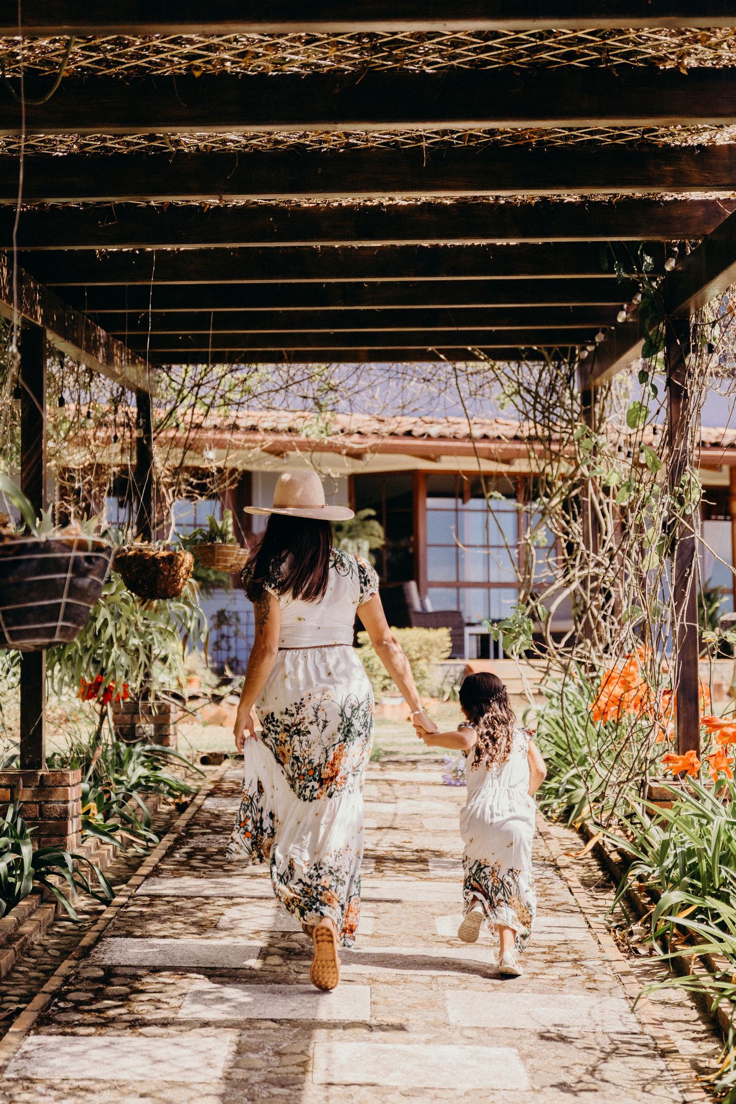 A mother and daughter in matching floral skirts walk hand in hand along a sunlit garden path.