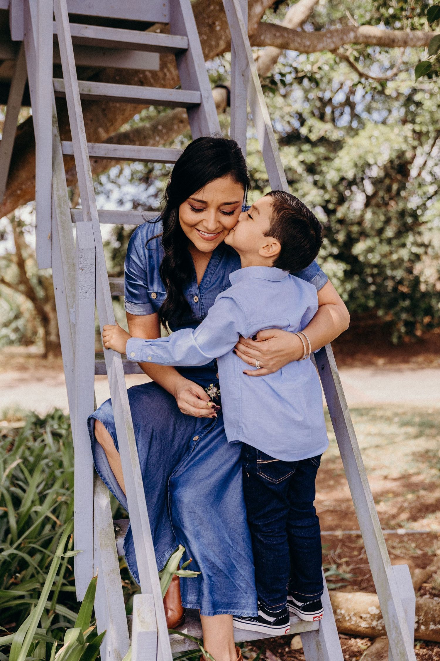 A young boy kisses his smiling mother on the cheek while they sit together on wooden steps in a lush garden.
