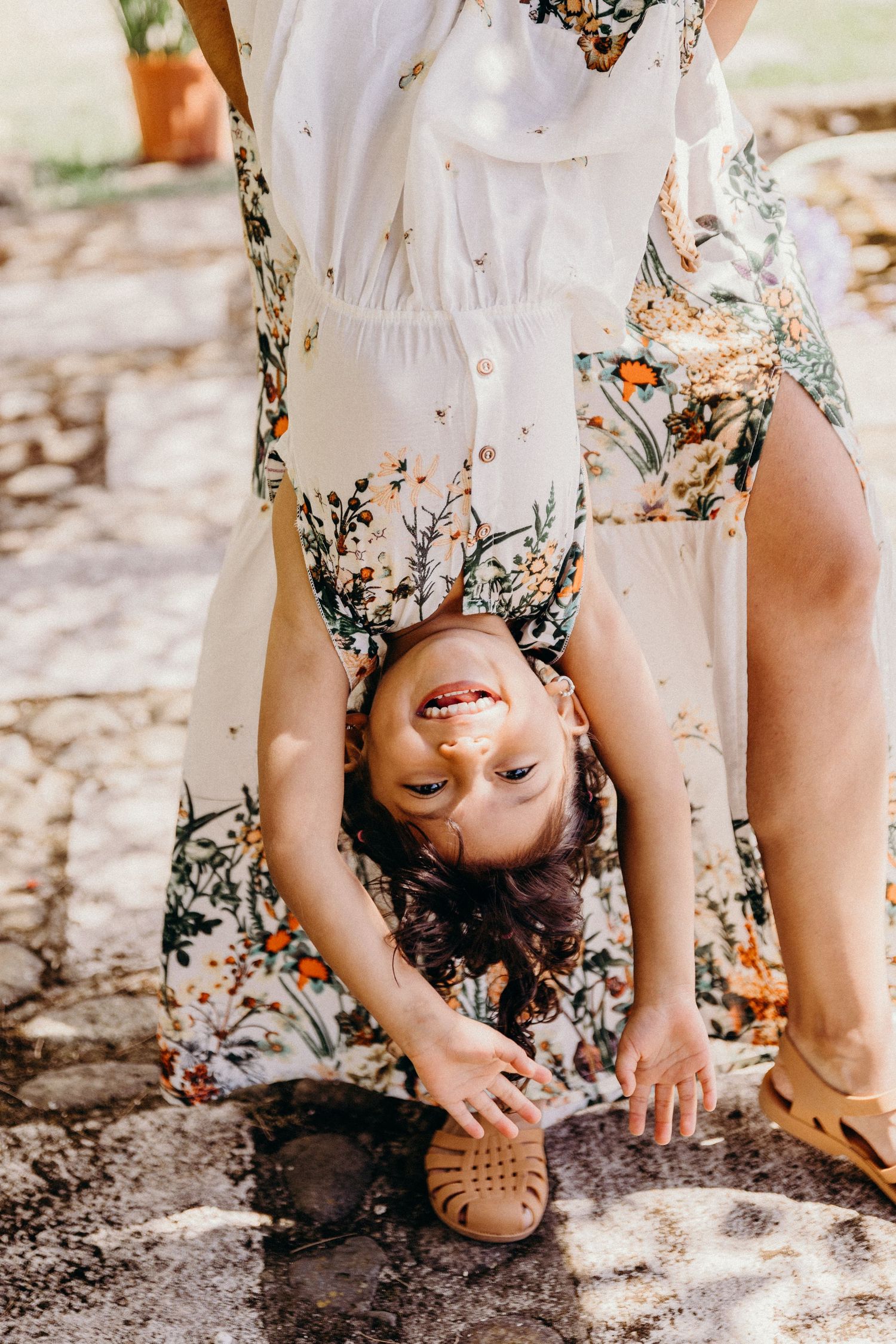 A laughing child held upside down by a woman in a floral dress outdoors in a sunny garden setting.