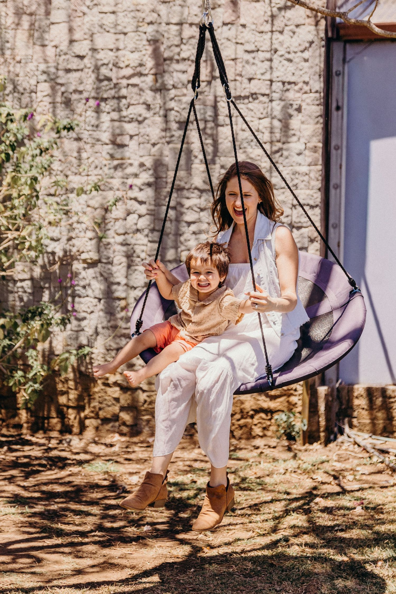 A smiling mother and young child joyfully sitting together on an outdoor hanging swing chair in a sunny garden.