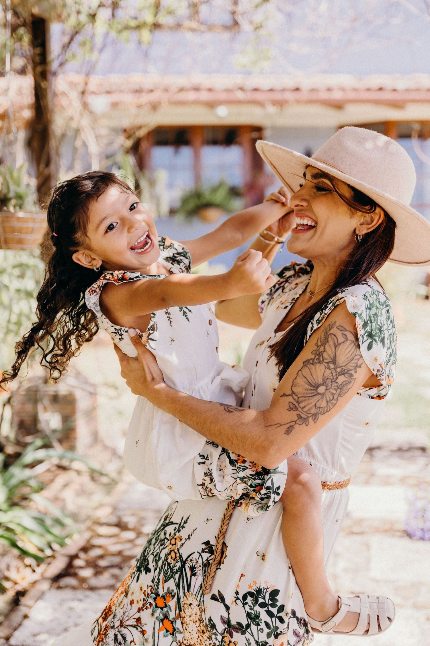 Mother and daughter in matching floral dresses laugh together outdoors, woman wearing hat and sunglasses with floral tattoo.