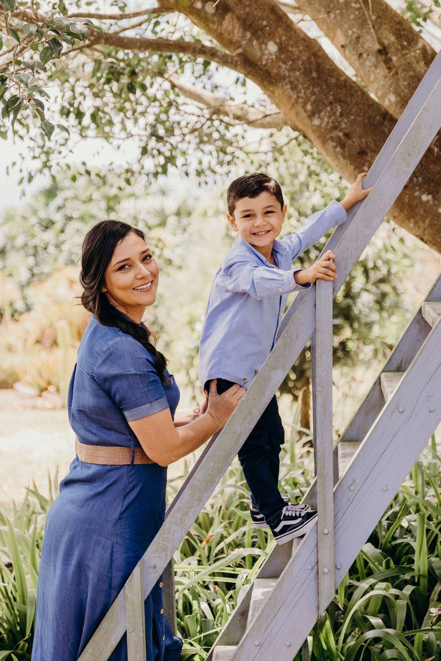 A woman in a blue dress and young boy in a light blue shirt smile on wooden stairs outdoors near a tree.