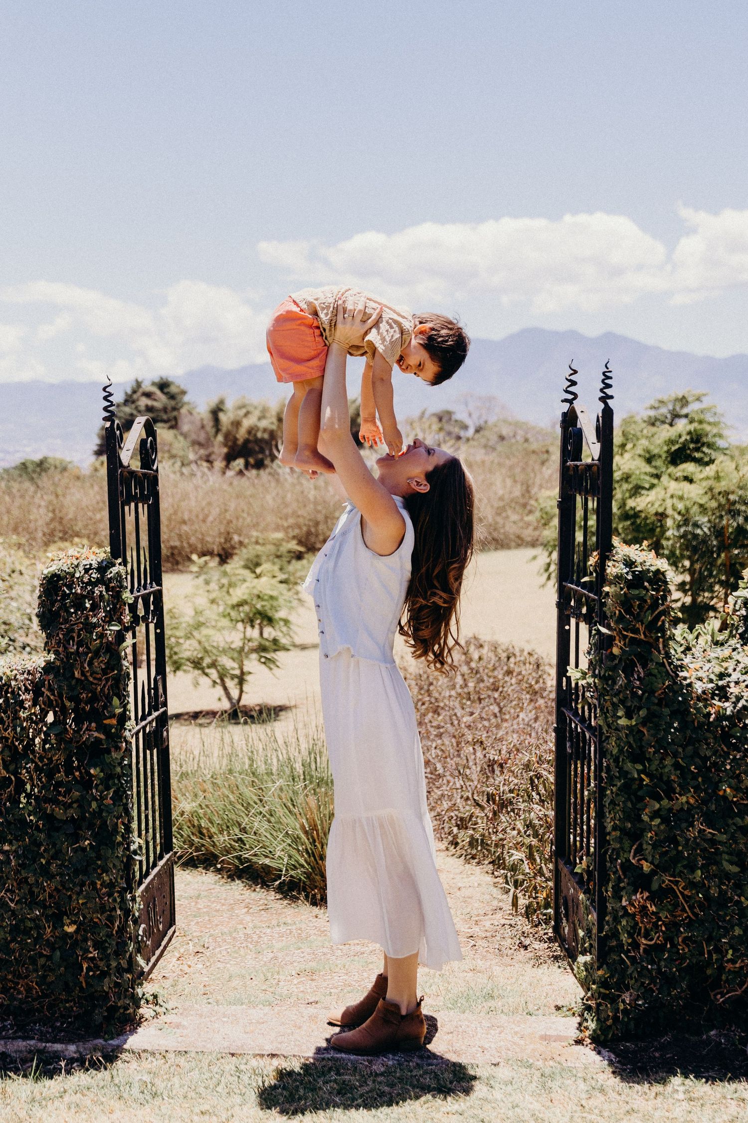 A mother in a white dress joyfully lifts her toddler overhead in a garden gate setting with mountains behind.