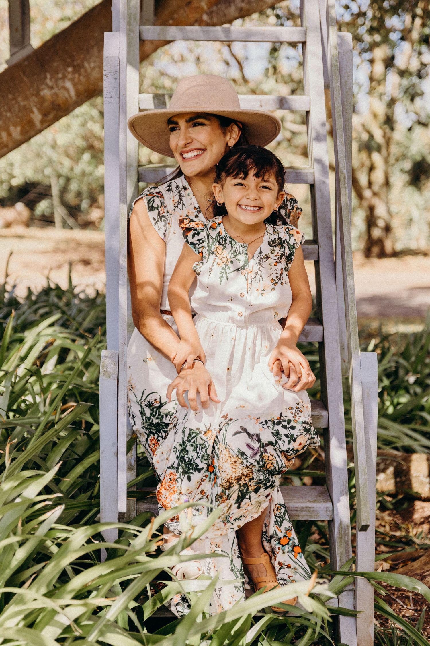 Smiling mother in beige hat and daughter wear matching floral dresses while sitting on wooden ladder steps outdoors.