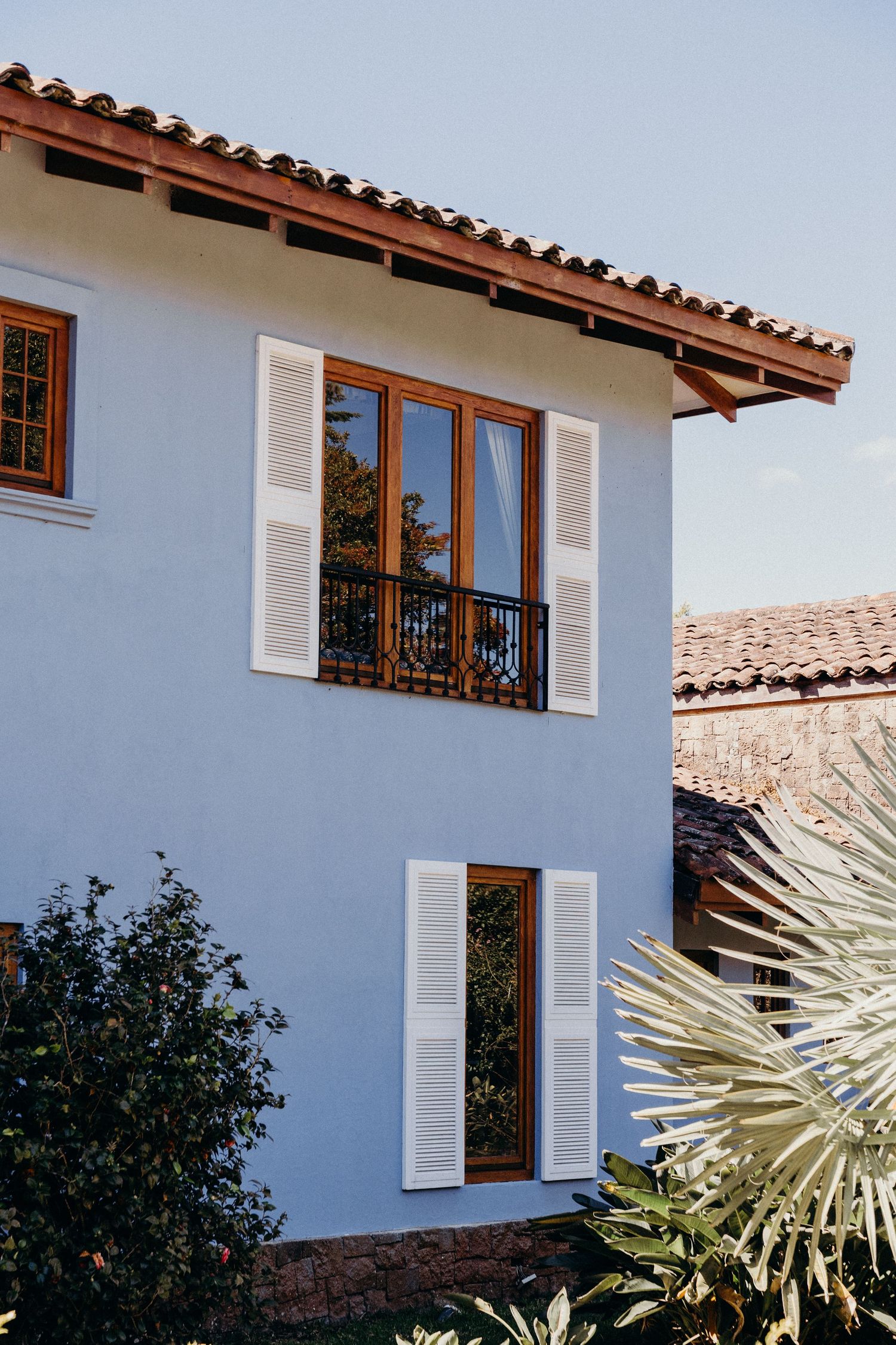 White stucco Mediterranean villa with terracotta roof, wooden shutters, and balcony surrounded by tropical plants.