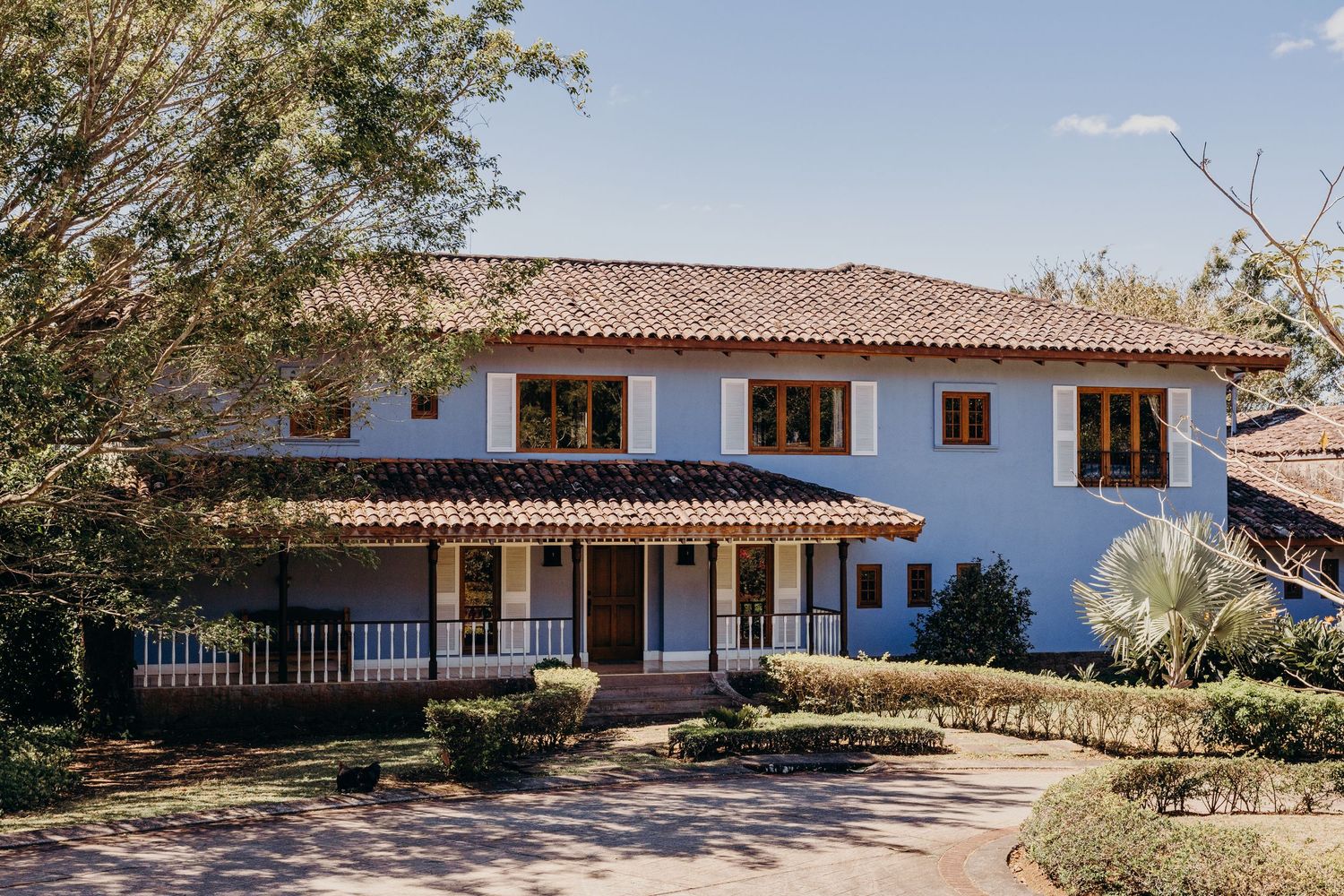 White Spanish colonial two-story house with terracotta roof, wooden railings, and manicured gardens on sunny day.