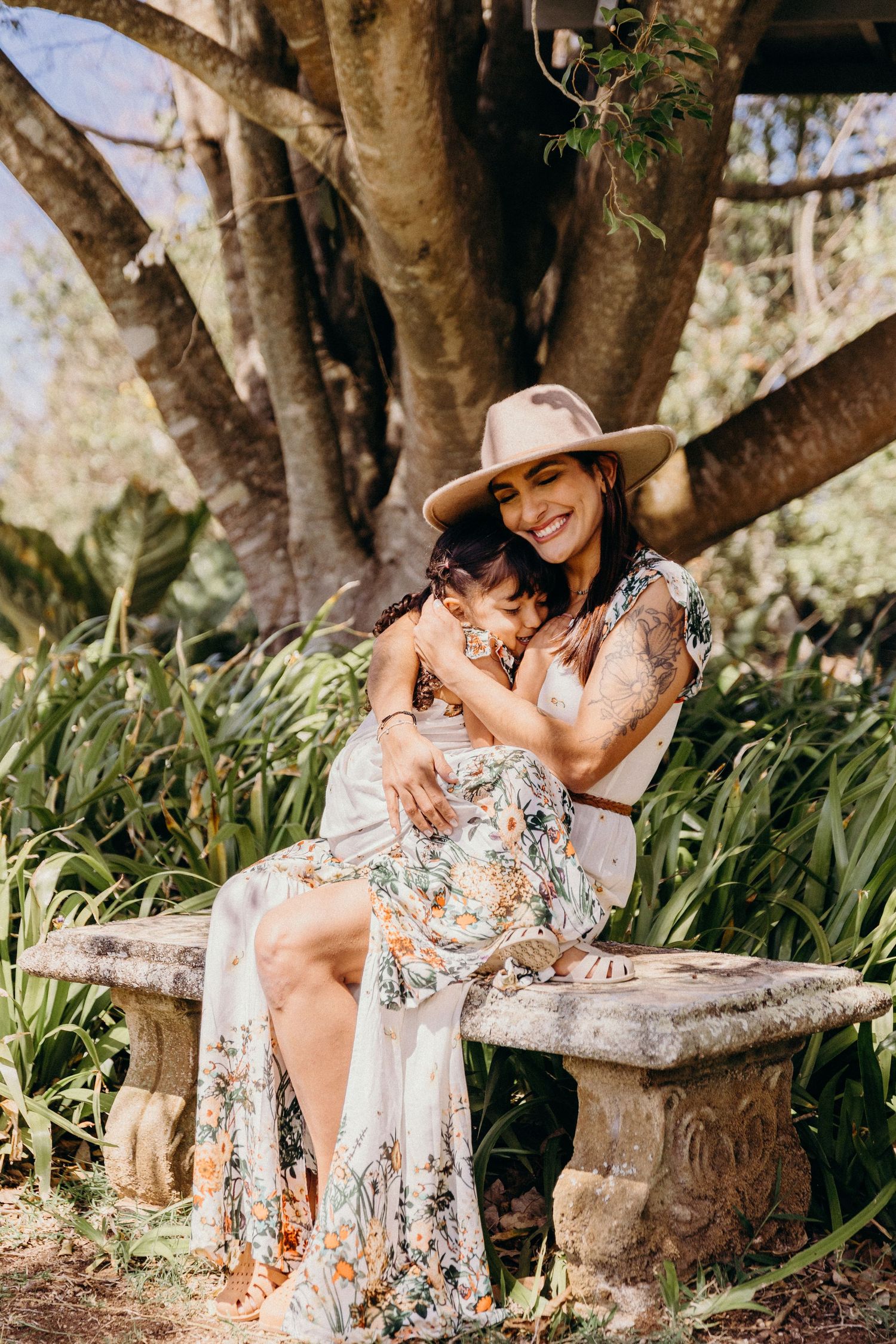 Woman in floral dress and hat hugs child on stone bench in lush garden setting.