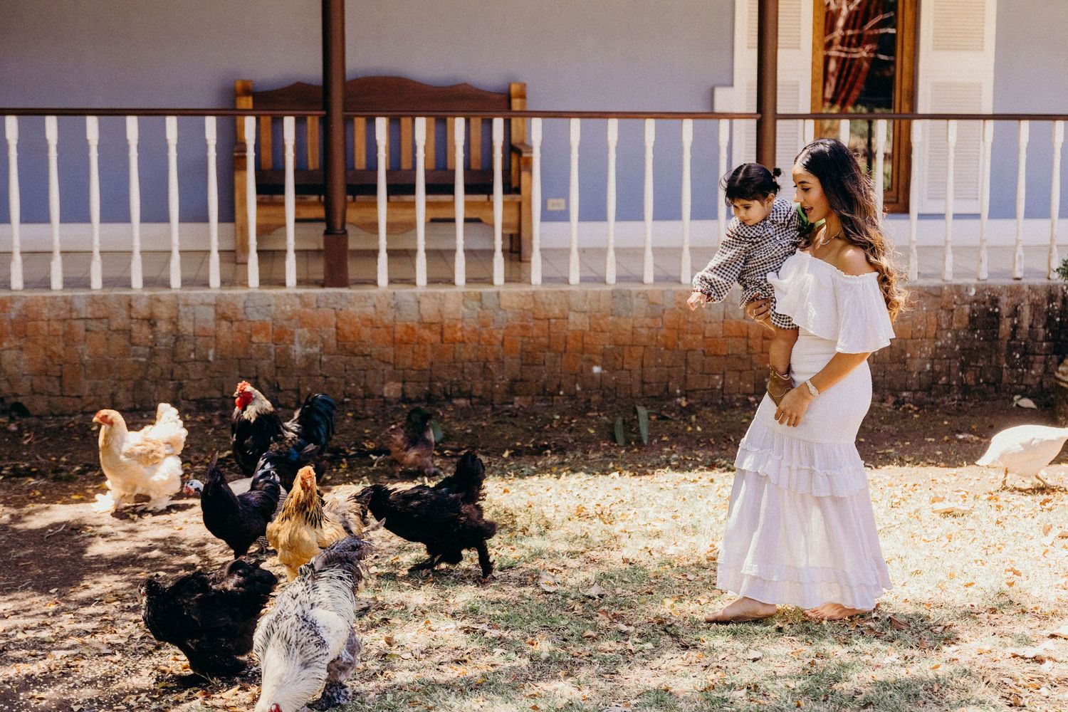 Woman in white dress holds baby while chickens roam freely in a rustic farmyard setting.