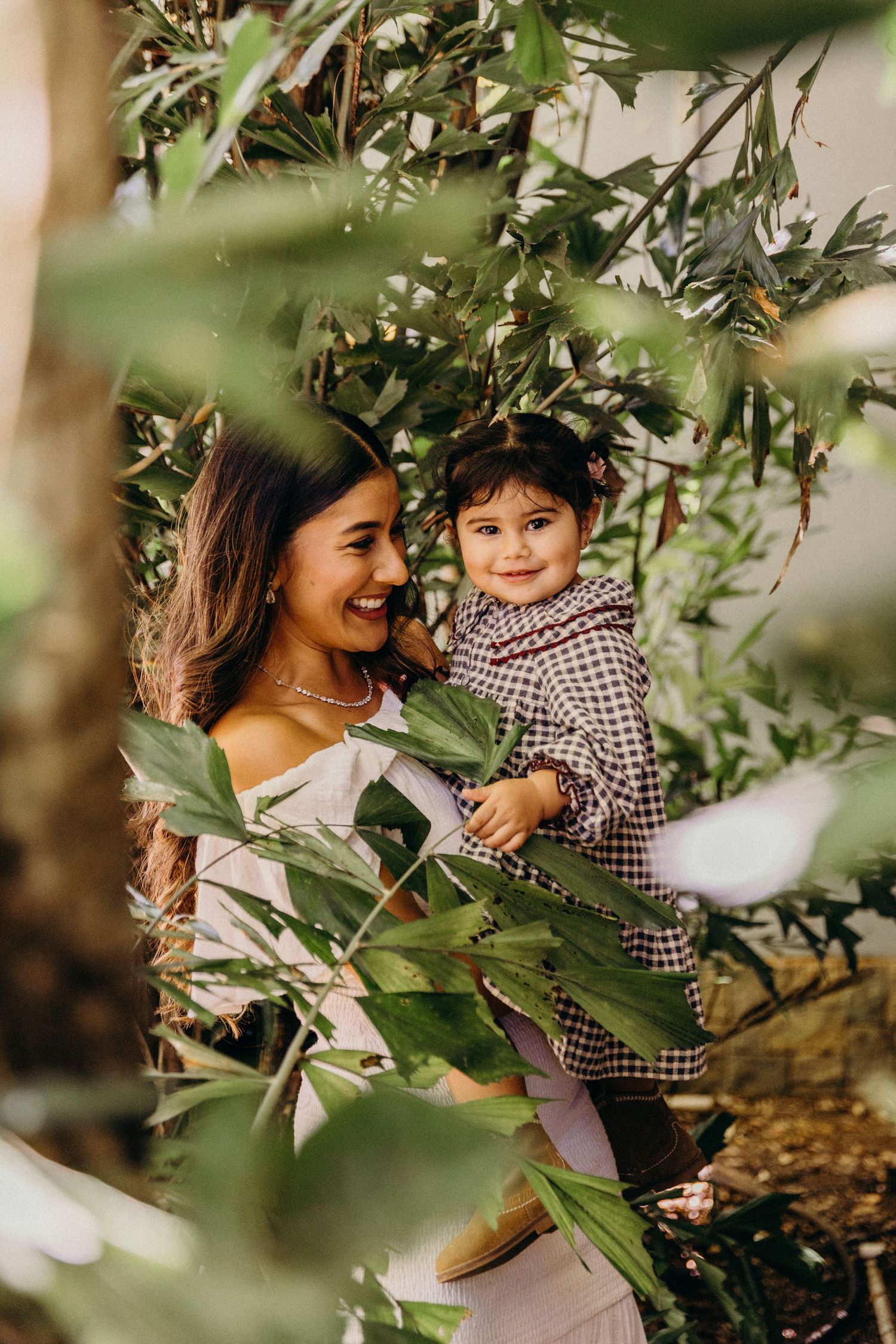 A smiling woman holds a toddler in a checkered outfit among lush green leaves in a garden setting.