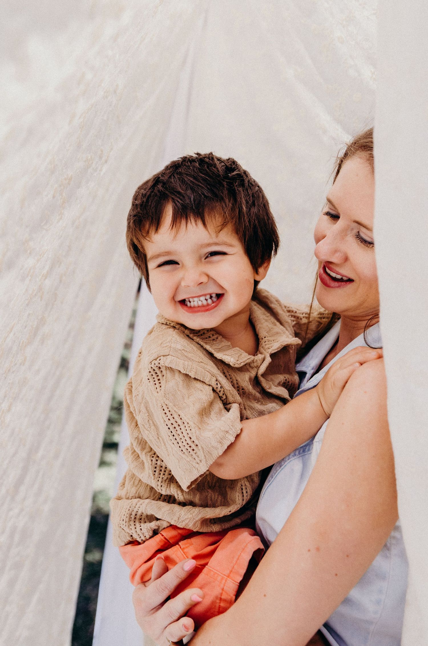 A joyful young boy smiles broadly while being held by a woman in a light setting with white drapes.