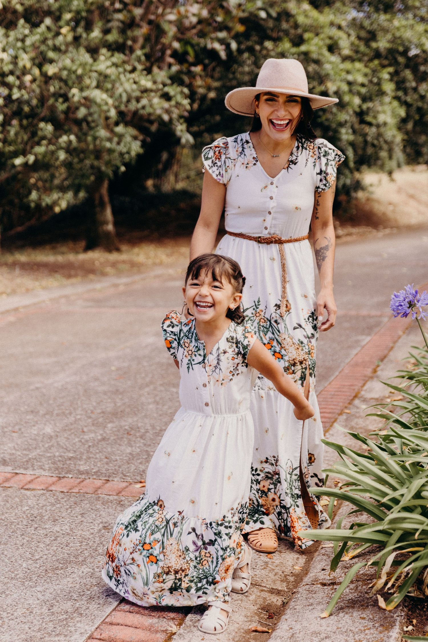 A laughing mother and daughter in matching white floral maxi dresses pose outdoors on a garden path.