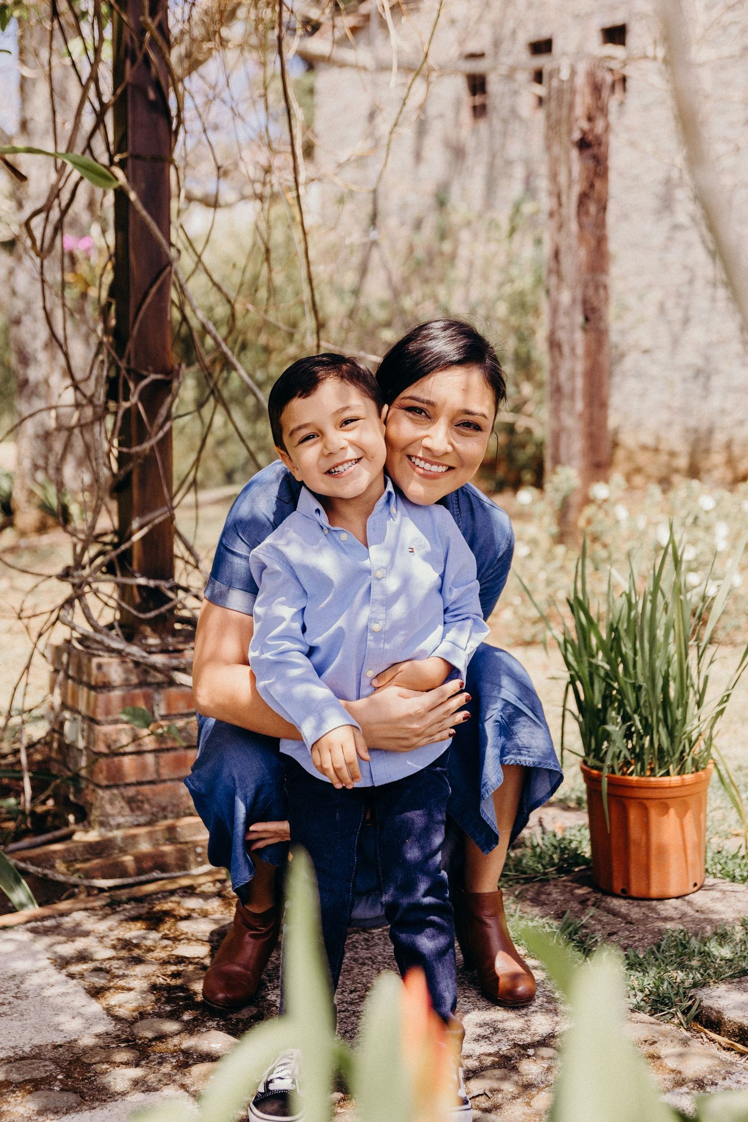 A smiling woman hugs a young boy in a sunny garden, both wearing blue outfits, sharing a joyful moment outdoors.