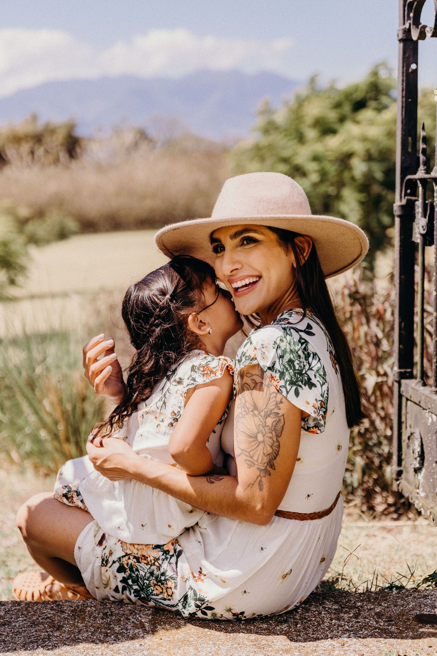 Woman in floral dress and hat hugging young girl outdoors with mountains in background.