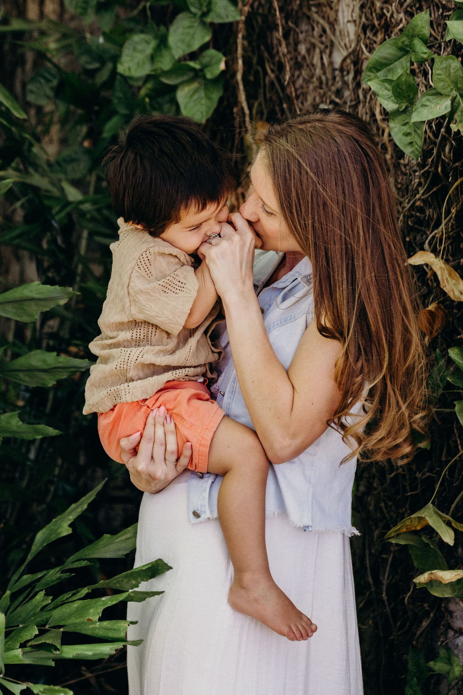 A mother tenderly holds her young son, touching noses and smiling, surrounded by lush green foliage.