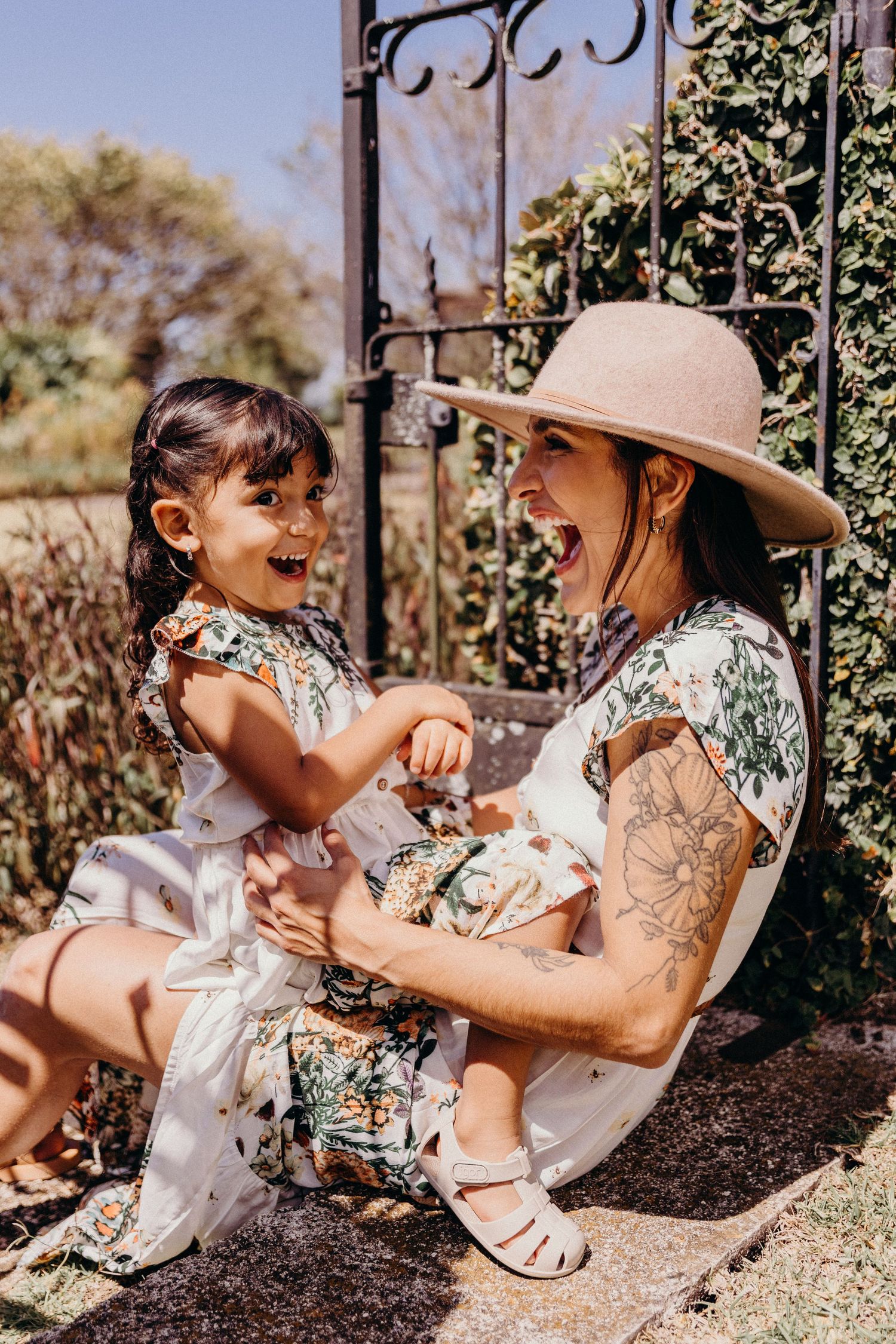 A laughing mother in a floral dress and hat plays with her joyful toddler daughter outdoors near a garden gate.