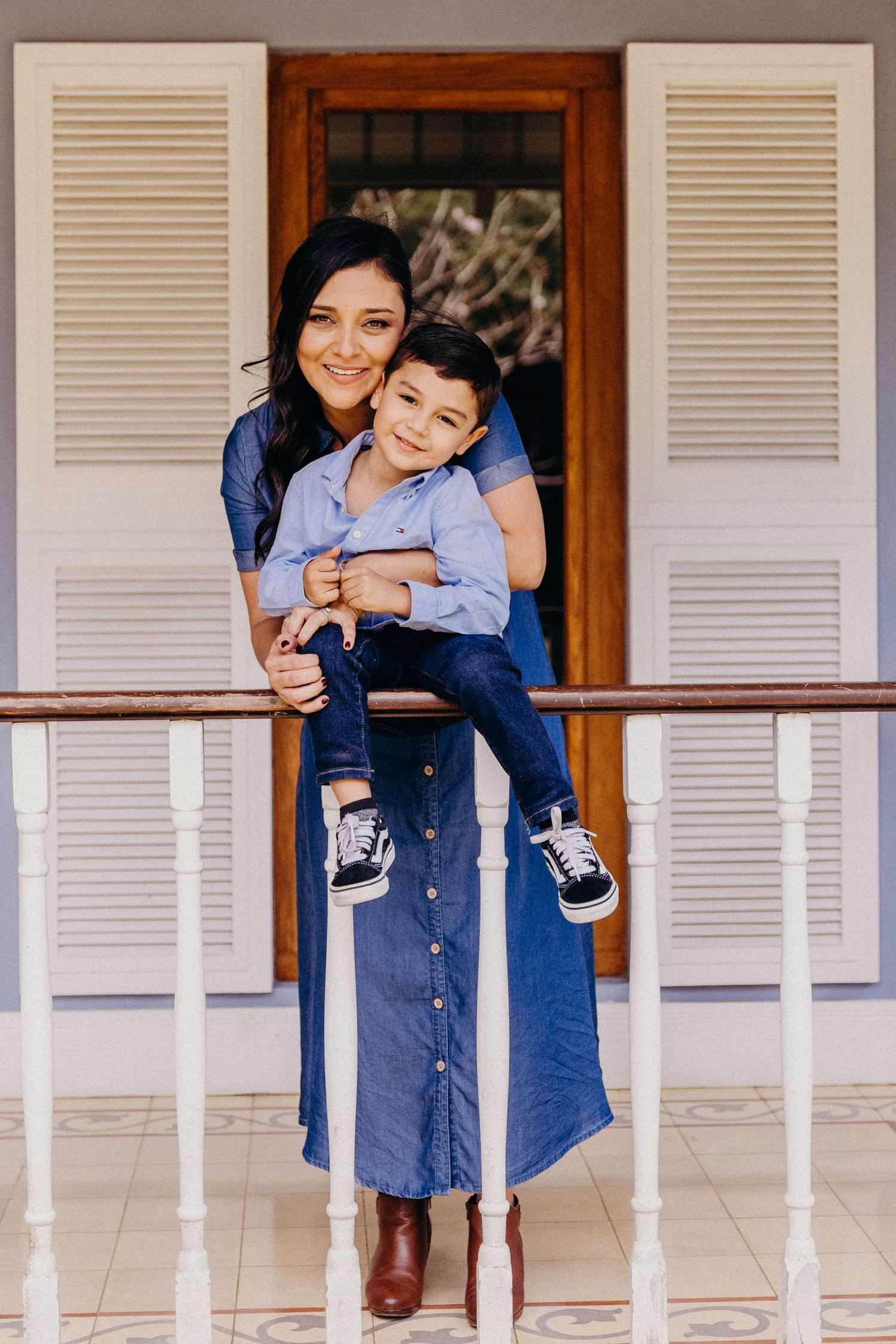 A woman in a denim dress holds a young boy on a porch railing, both smiling warmly.