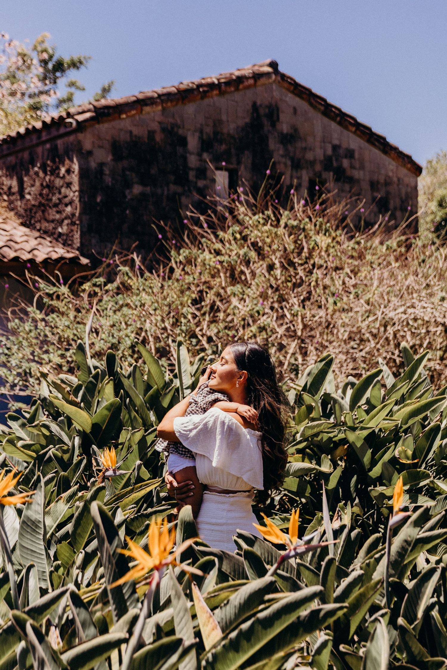 Woman in white dress standing among bird of paradise flowers with a rustic building in the background.