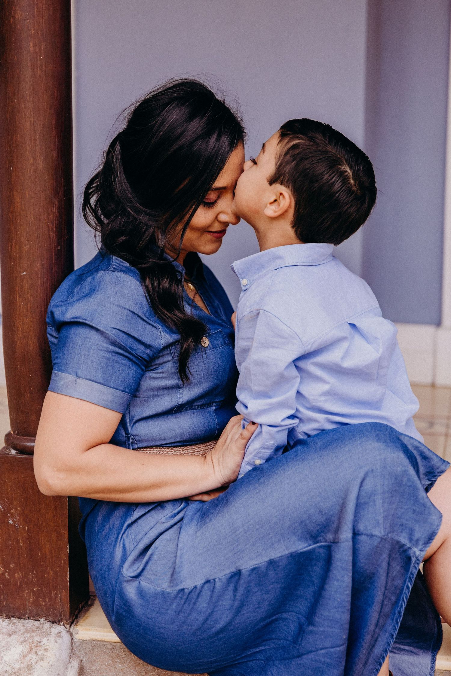 A mother in a blue dress shares a tender kiss with her young son sitting on her lap outdoors.