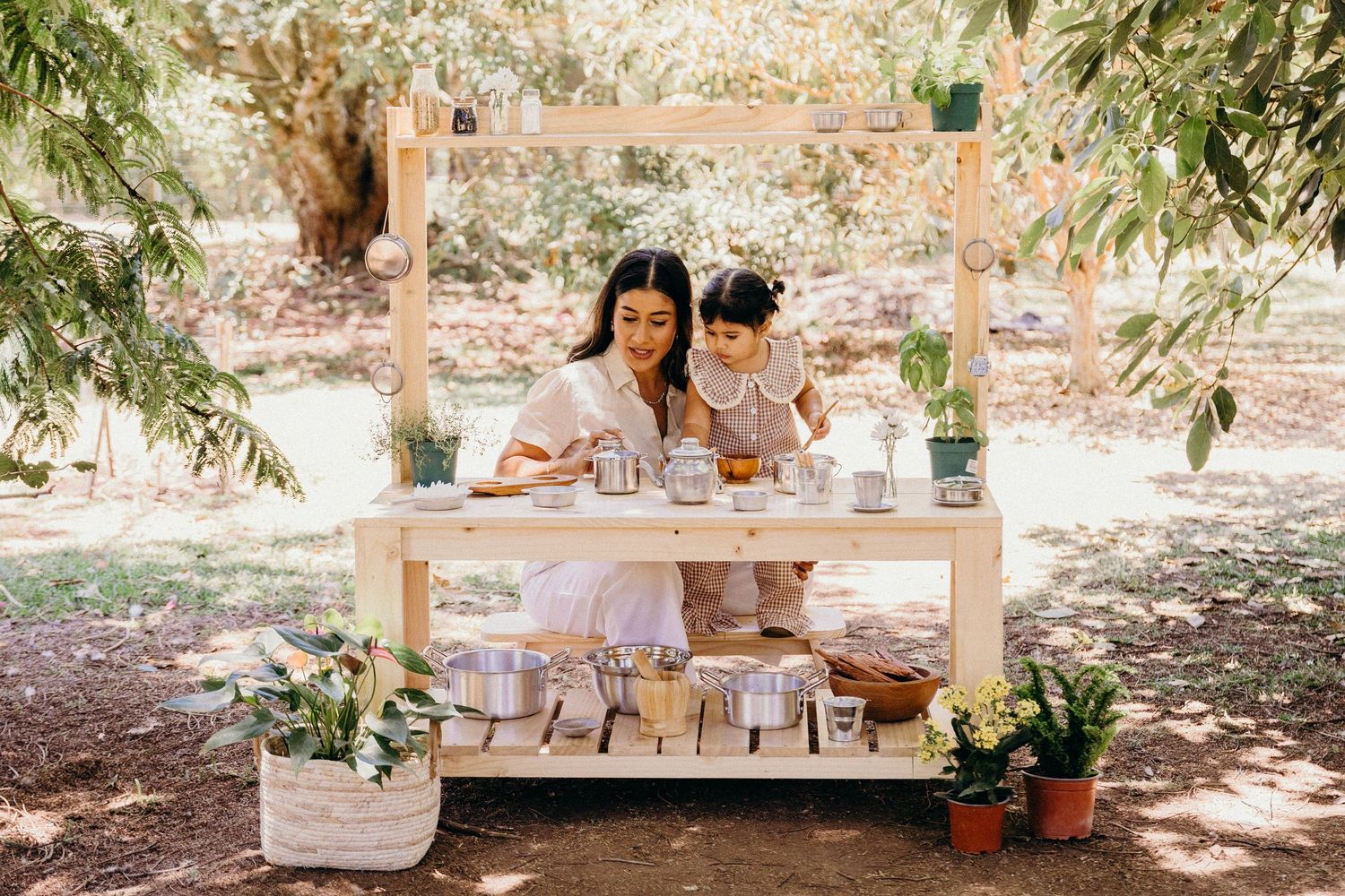 Woman and child explore a rustic wooden outdoor market stand surrounded by plants and natural decor in a serene garden setting.
