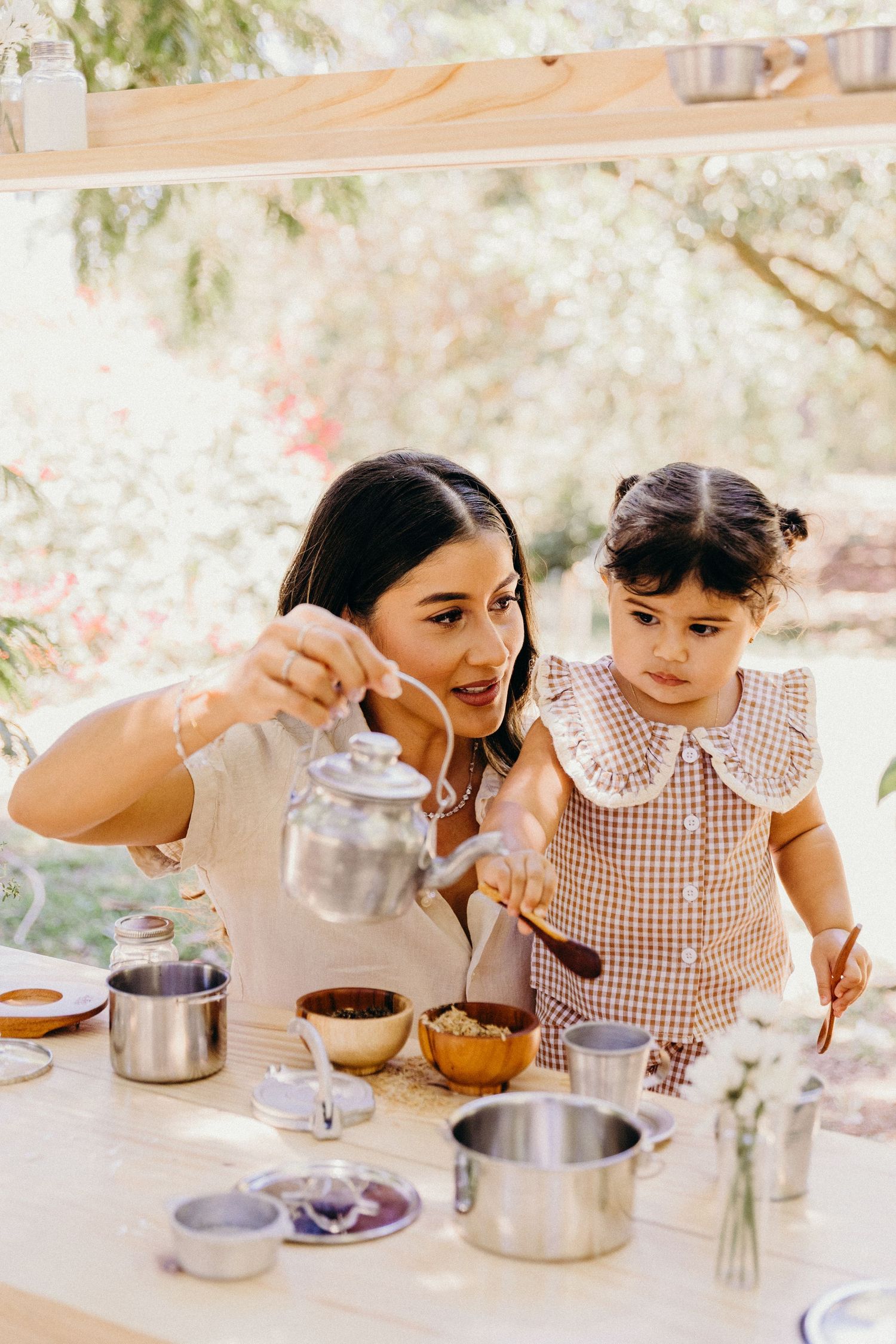 A mother and toddler girl play with a silver tea set at an outdoor table surrounded by lush greenery.