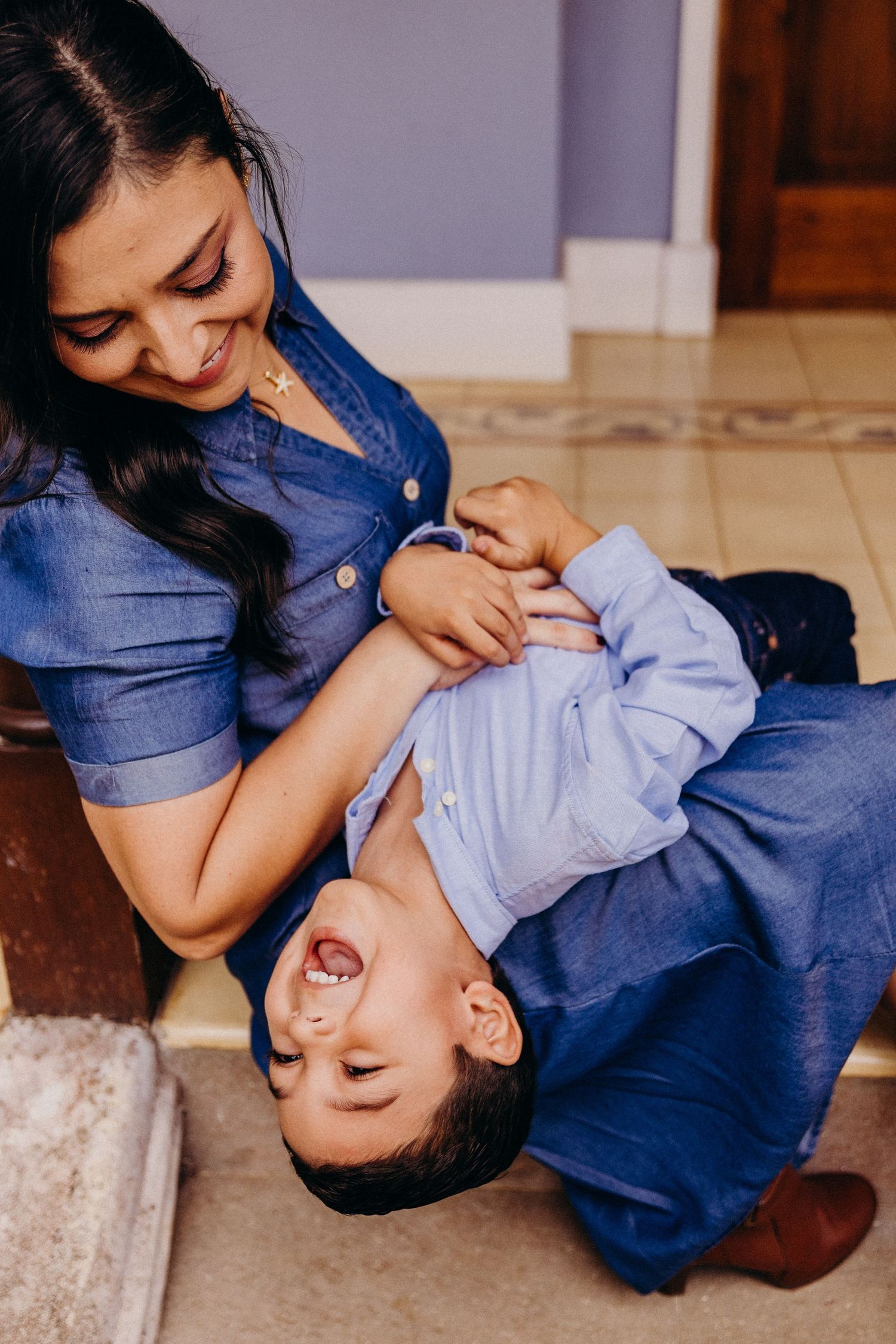 A smiling mother in a blue dress playfully holds her laughing baby boy on the floor indoors.