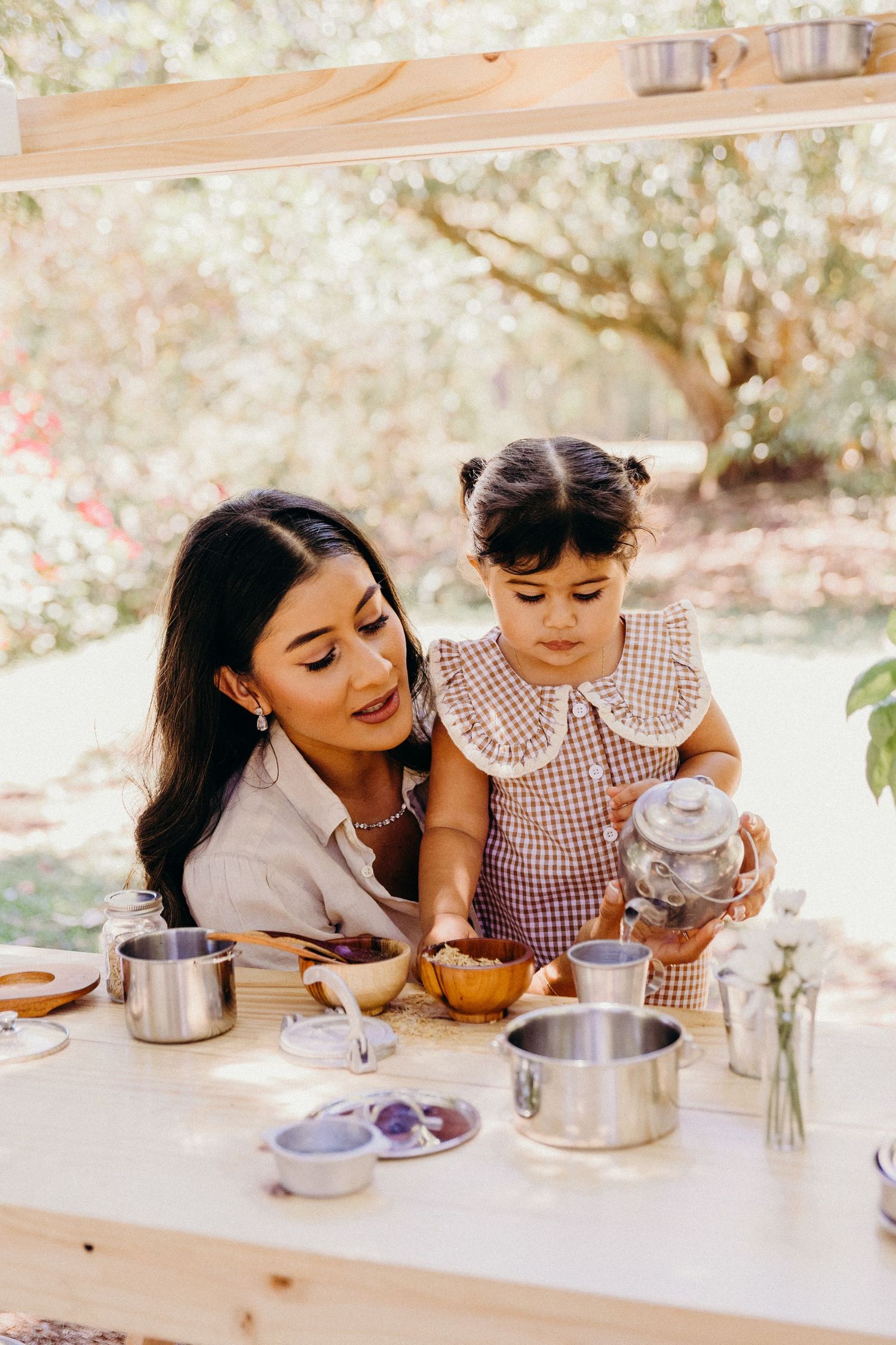 Mother and toddler girl playing with toy tea set at wooden outdoor kitchen stand surrounded by blooming garden.