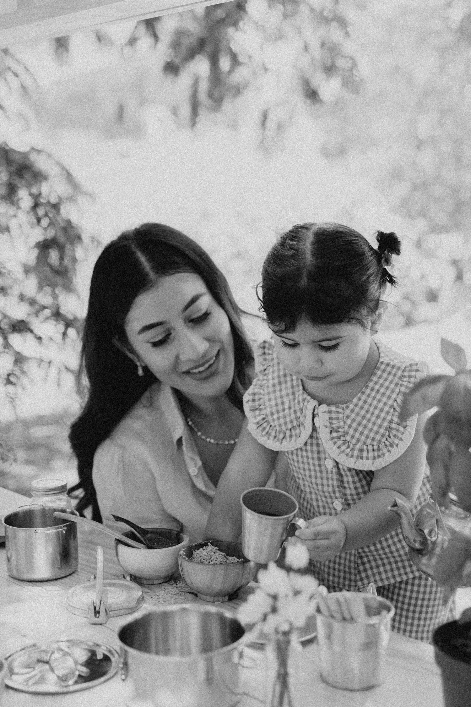 Black and white photo of a smiling mother and young daughter playing with metal cups and bowls at a table.