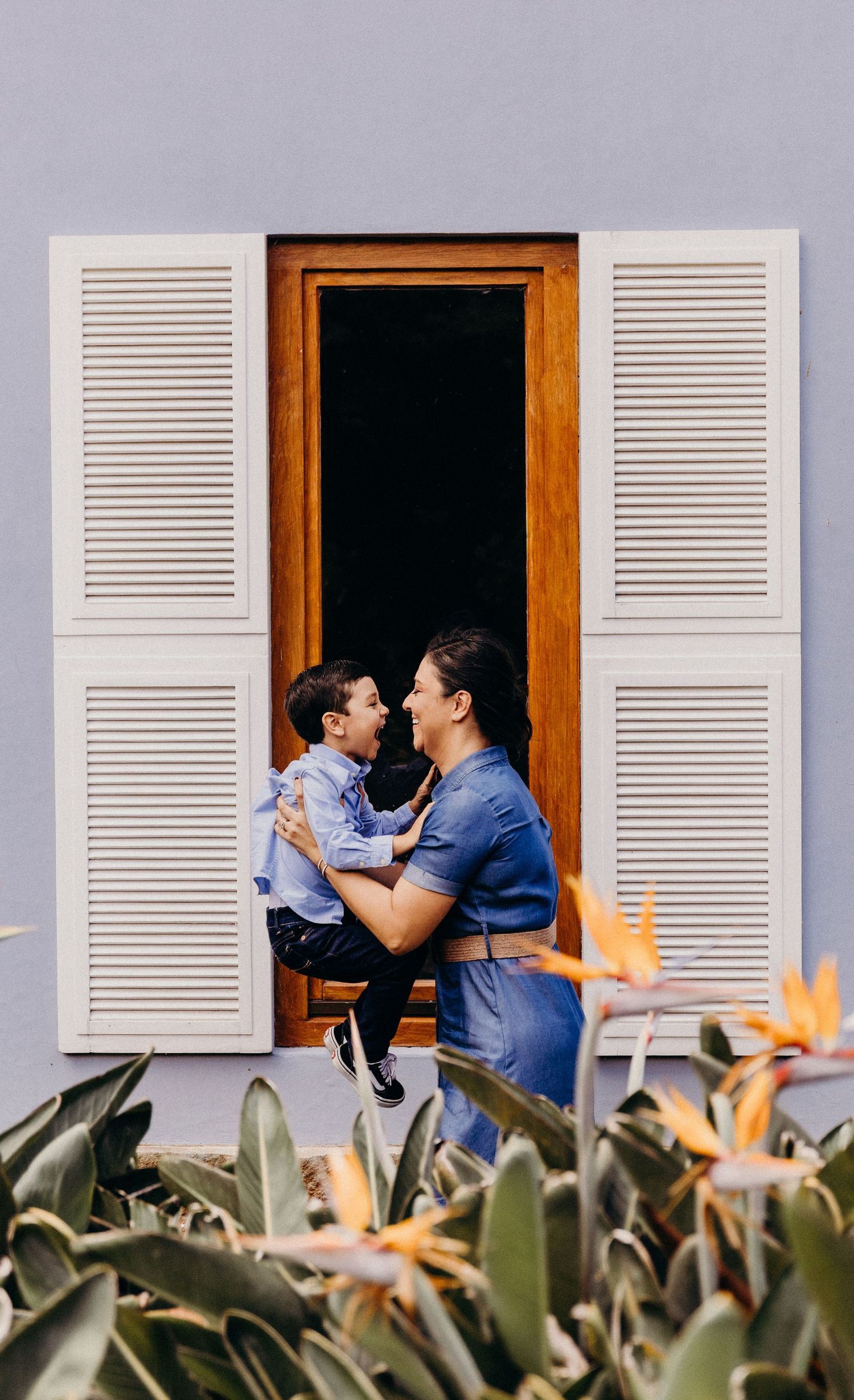 A mother holds her young son in a window with white shutters, surrounded by tropical plants and bird of paradise flowers.