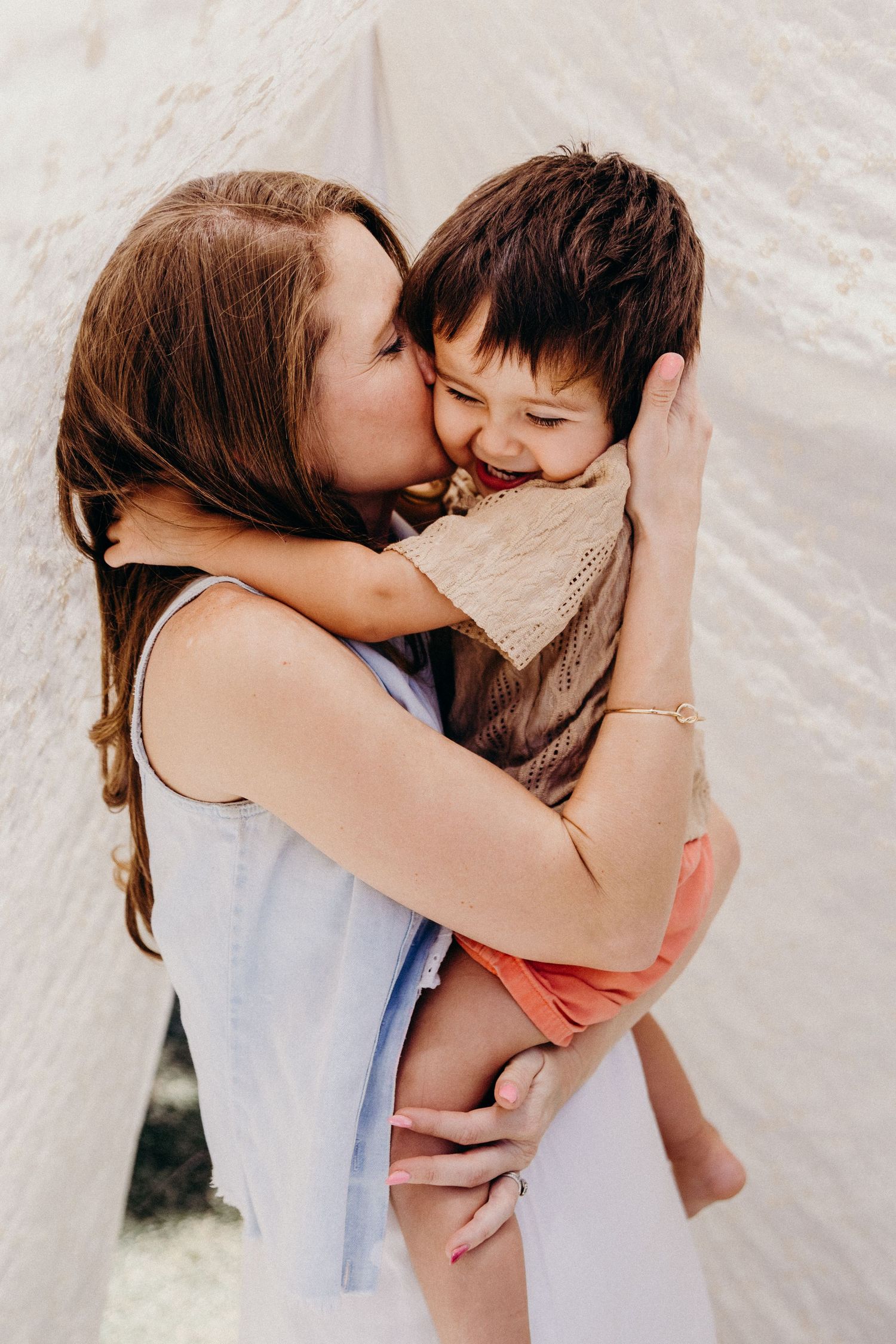 A mother tenderly kisses her young son on the cheek while holding him in her arms outdoors.