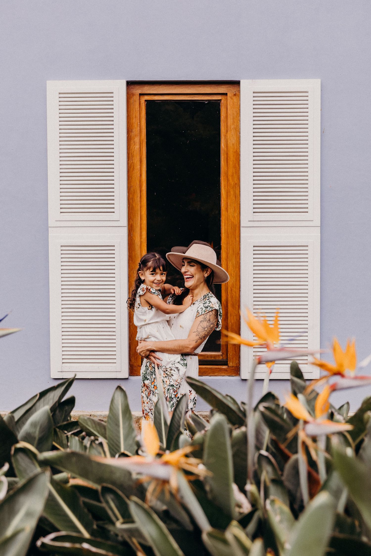 A smiling woman in a hat holds a young child in front of a blue wall with white shuttered window and tropical plants.