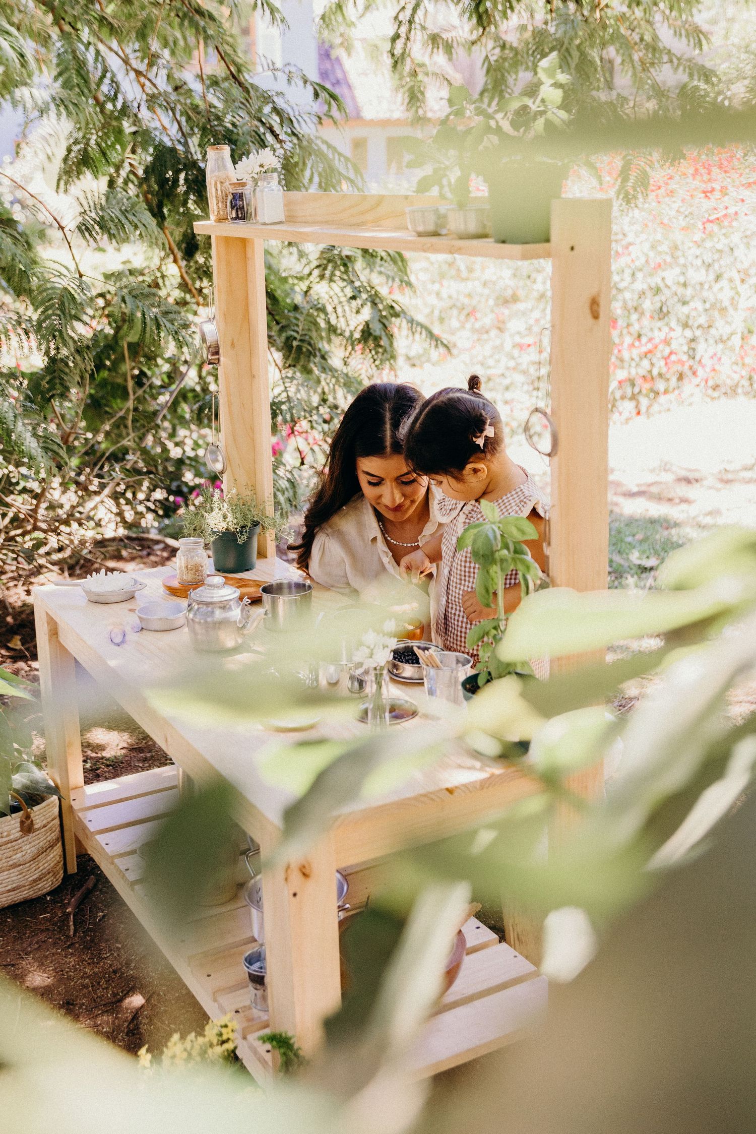 A woman and child play together at a wooden outdoor kitchen set surrounded by lush green garden foliage.