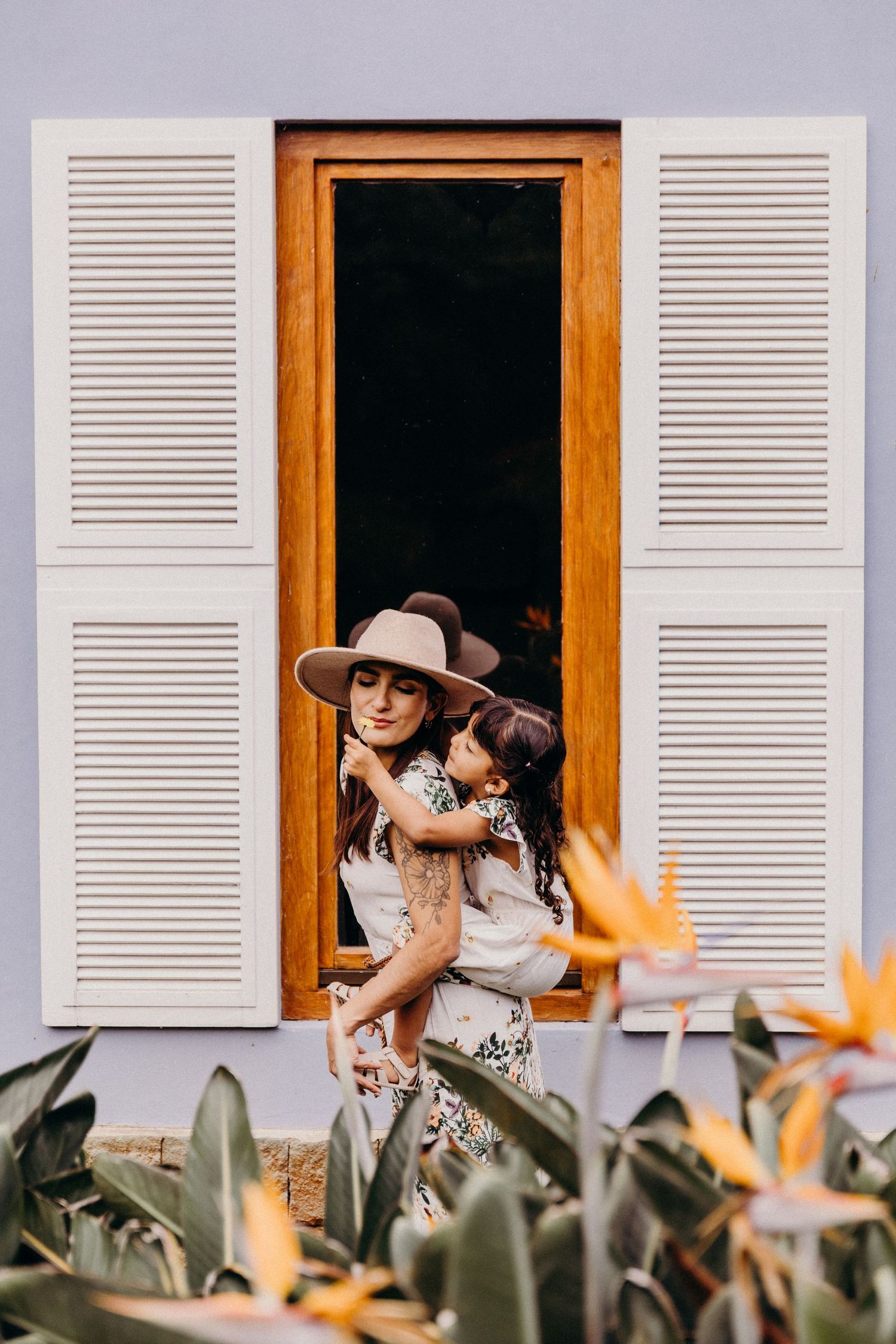 Woman in wide-brim hat holding a child in front of wooden-framed window with white shutters and tropical plants.