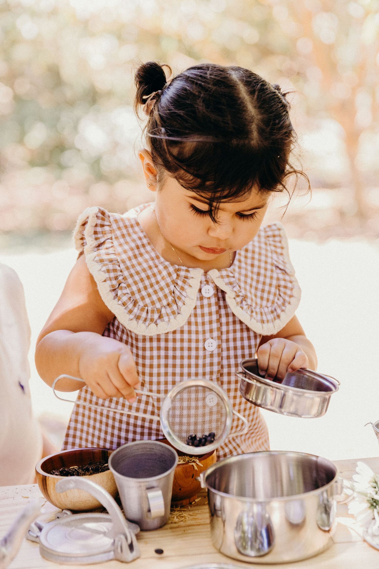 Toddler girl in gingham dress playing with miniature pots and pans at outdoor pretend cooking station.