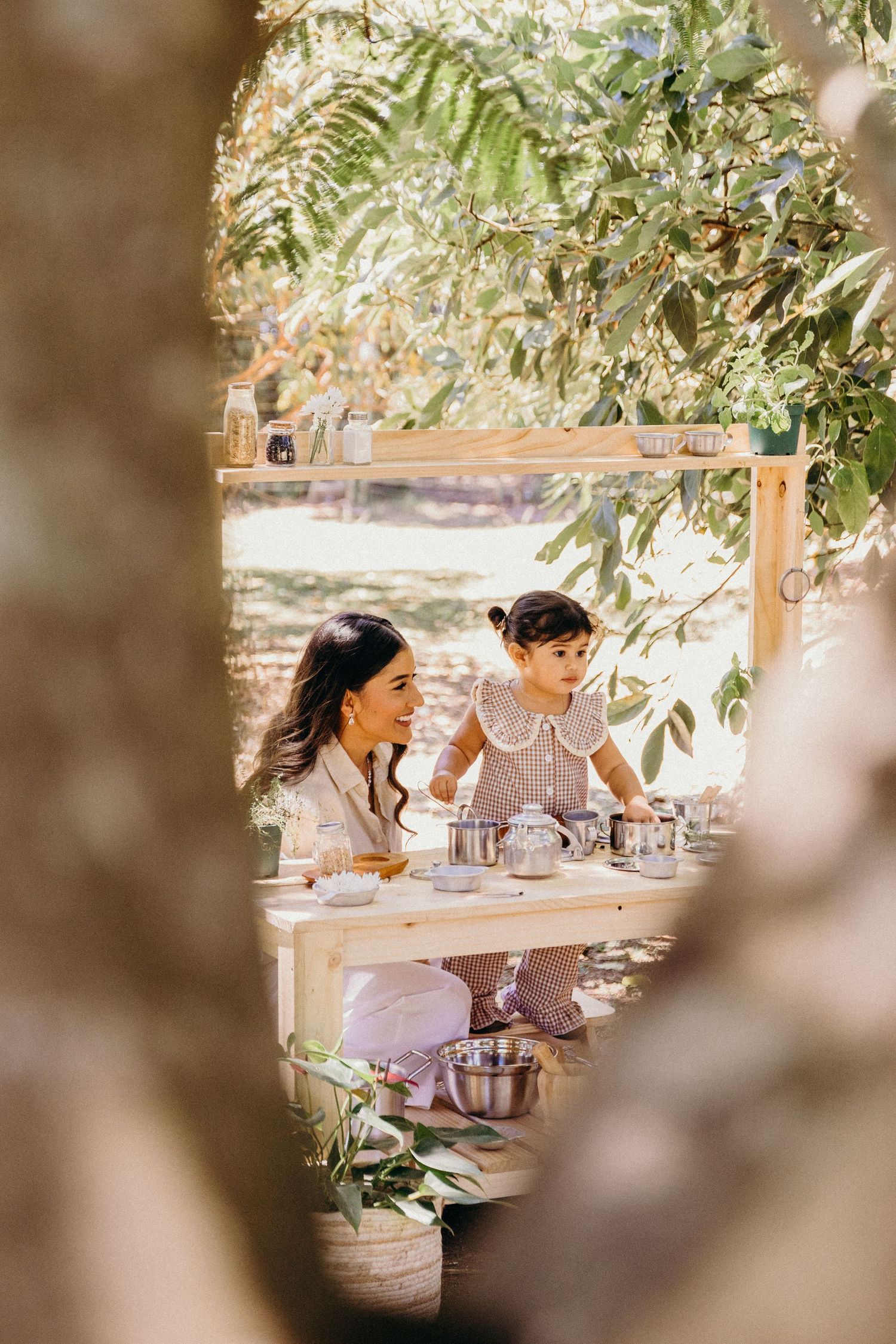 A mother and toddler enjoy an outdoor tea party at a wooden table surrounded by lush green trees.