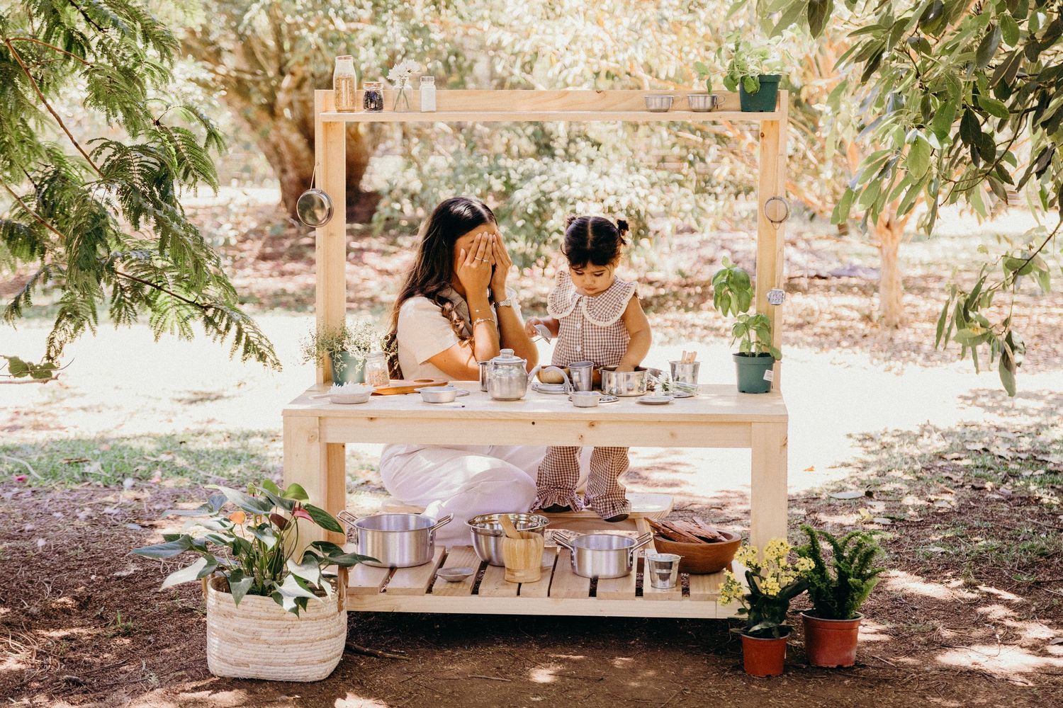 Woman and child at a rustic wooden market stall with plants and goods in a shaded outdoor garden setting.