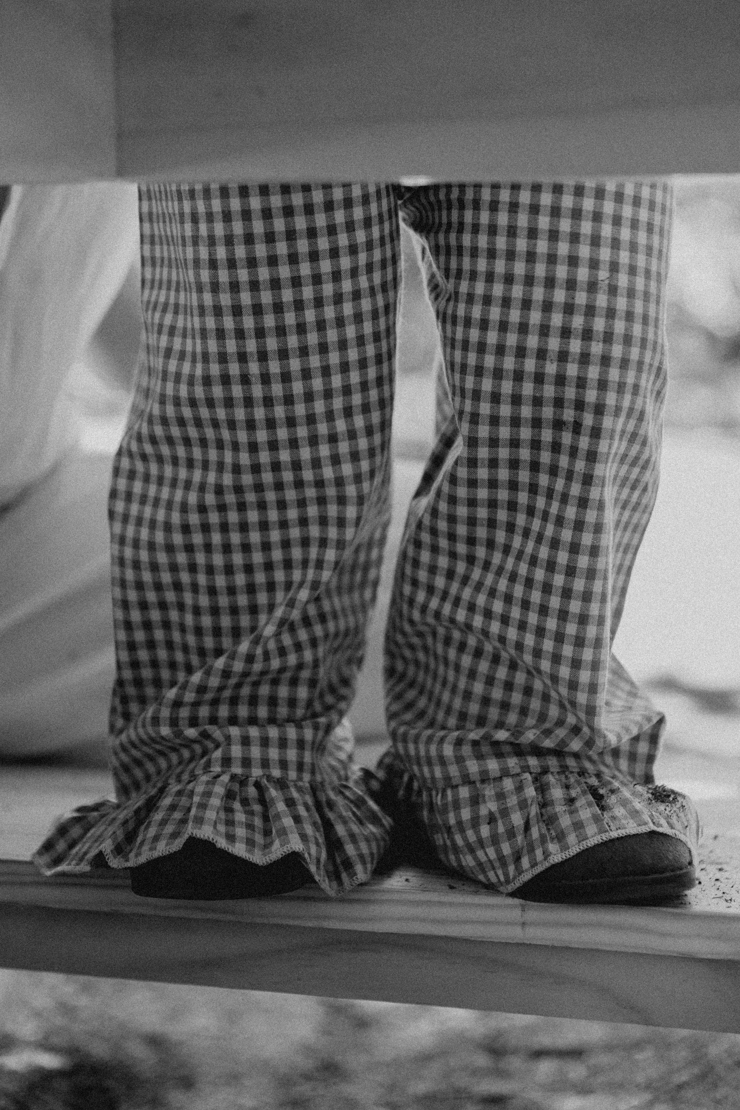 Black and white photo of child wearing ruffled gingham check pants standing on wooden bench.