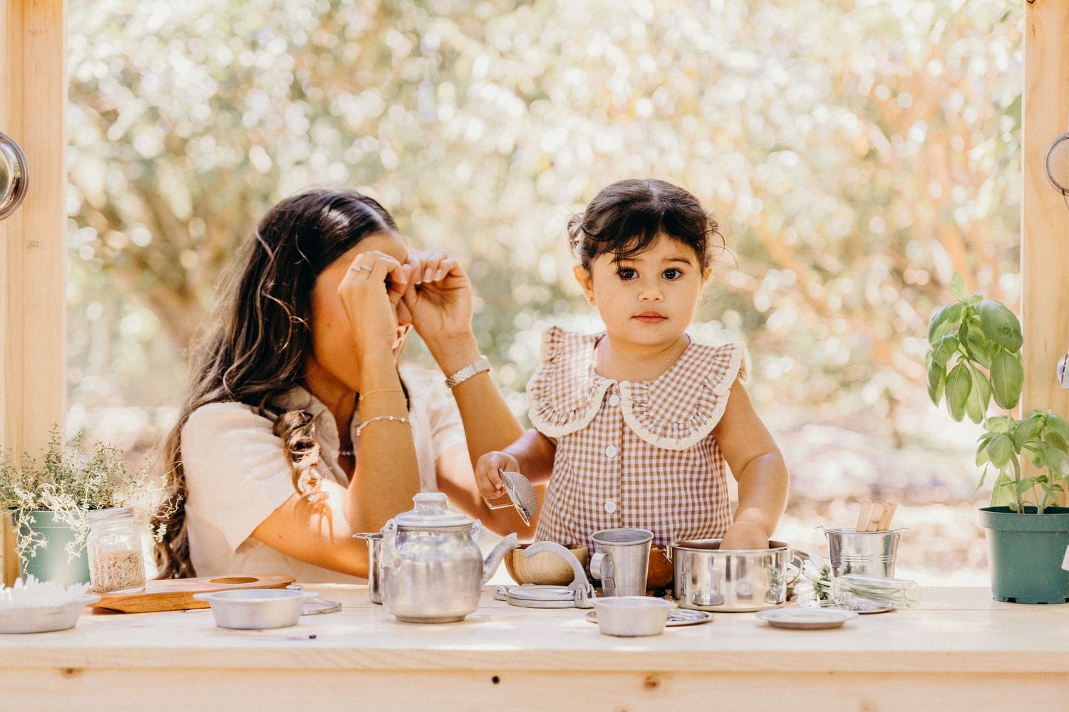 Two young girls playing tea party with silver tea set on wooden table near bright window.