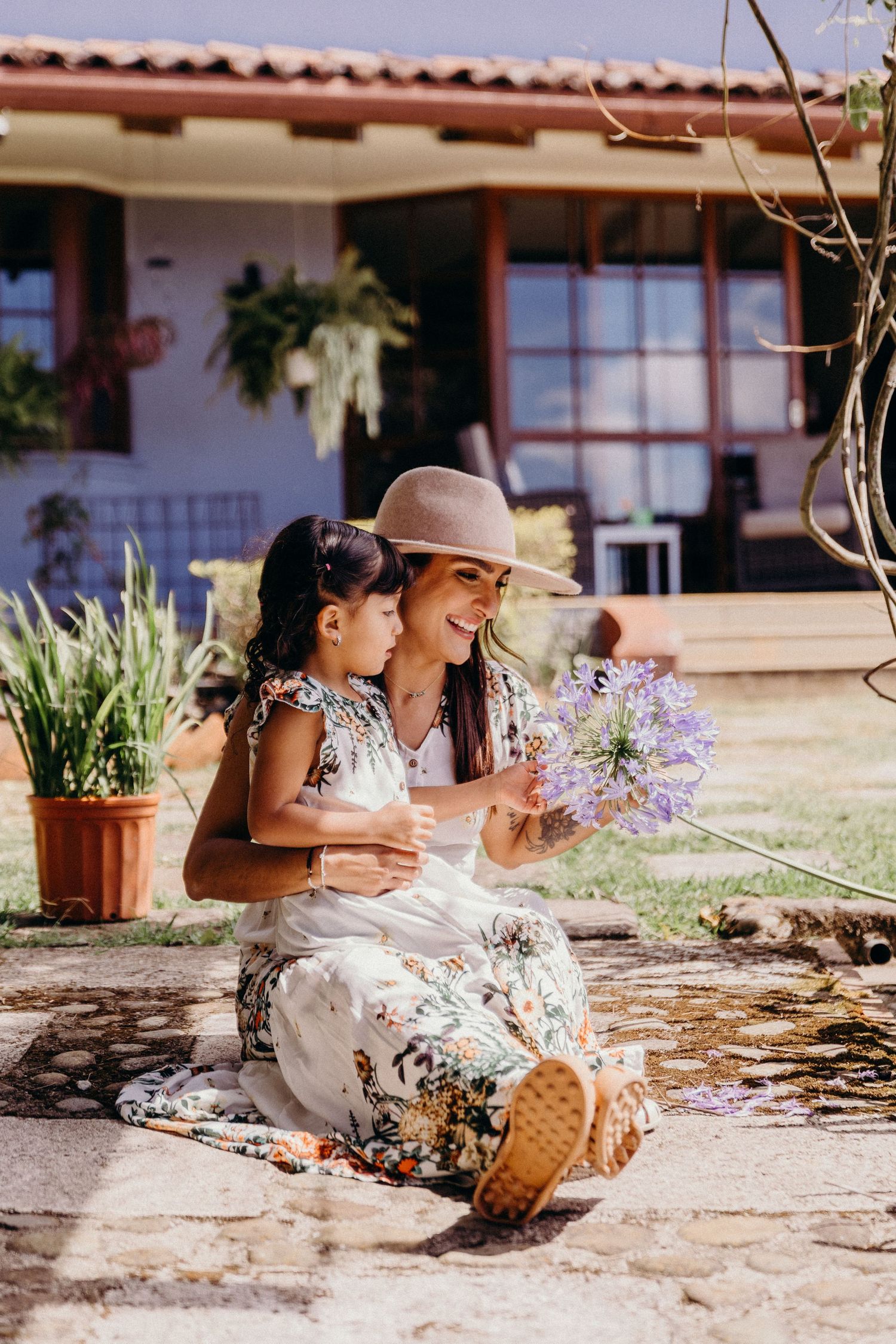 A woman in a floral dress and hat sits with a young girl, admiring purple flowers outside a home.