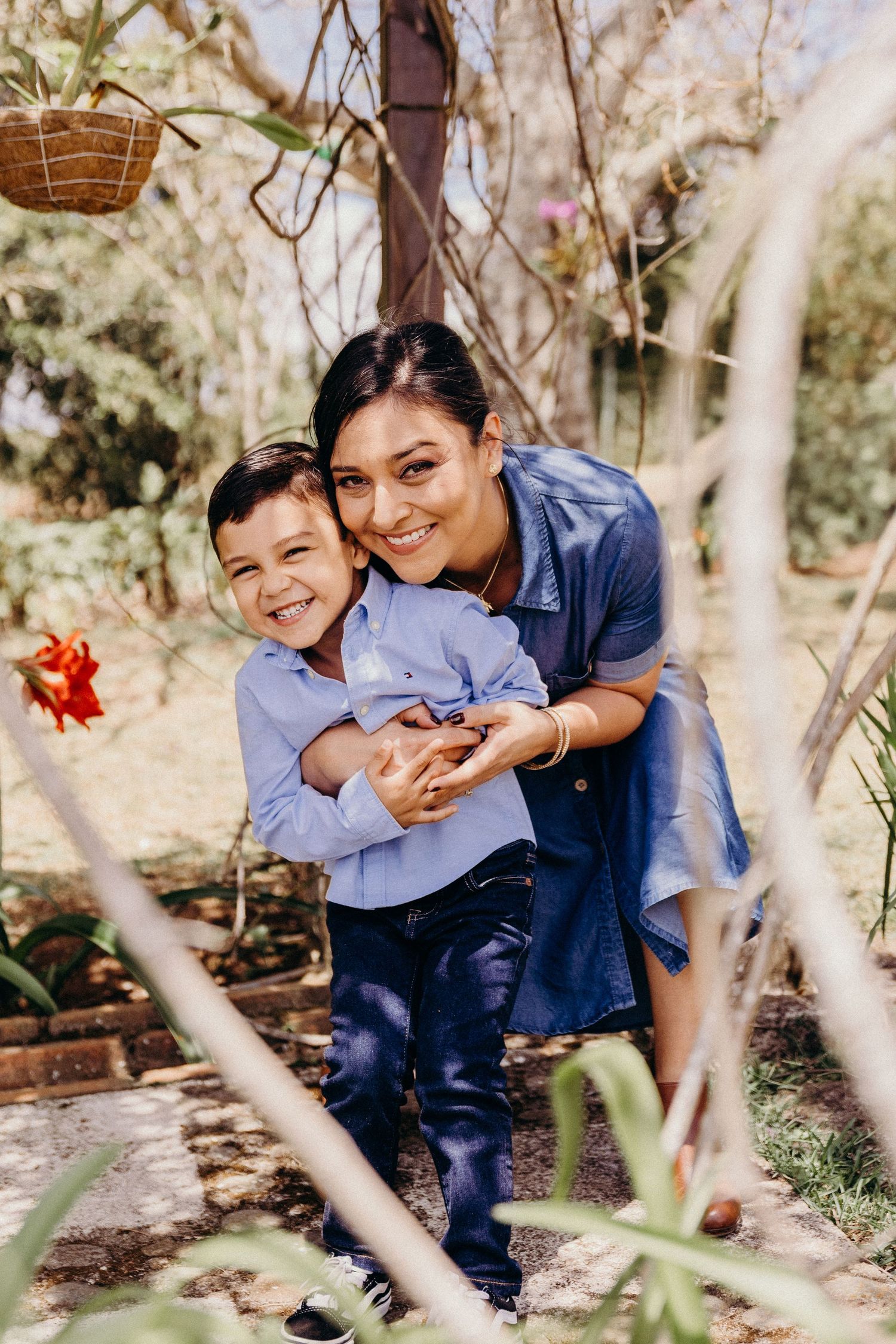 A smiling woman hugs a young boy from behind in a sunny garden, both wearing blue outfits.