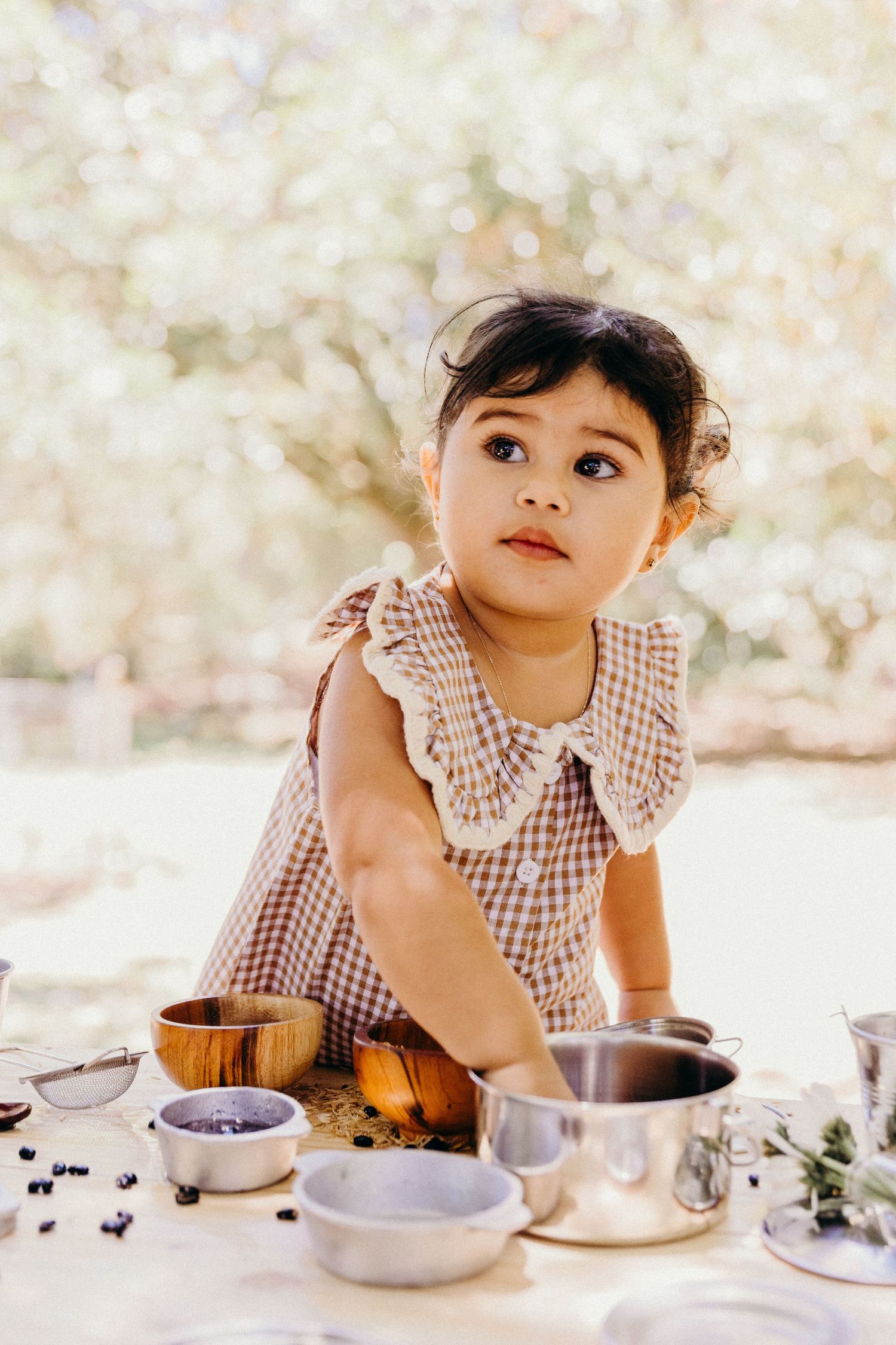 Toddler girl in gingham dress playing at outdoor table with bowls and blueberries in natural light.