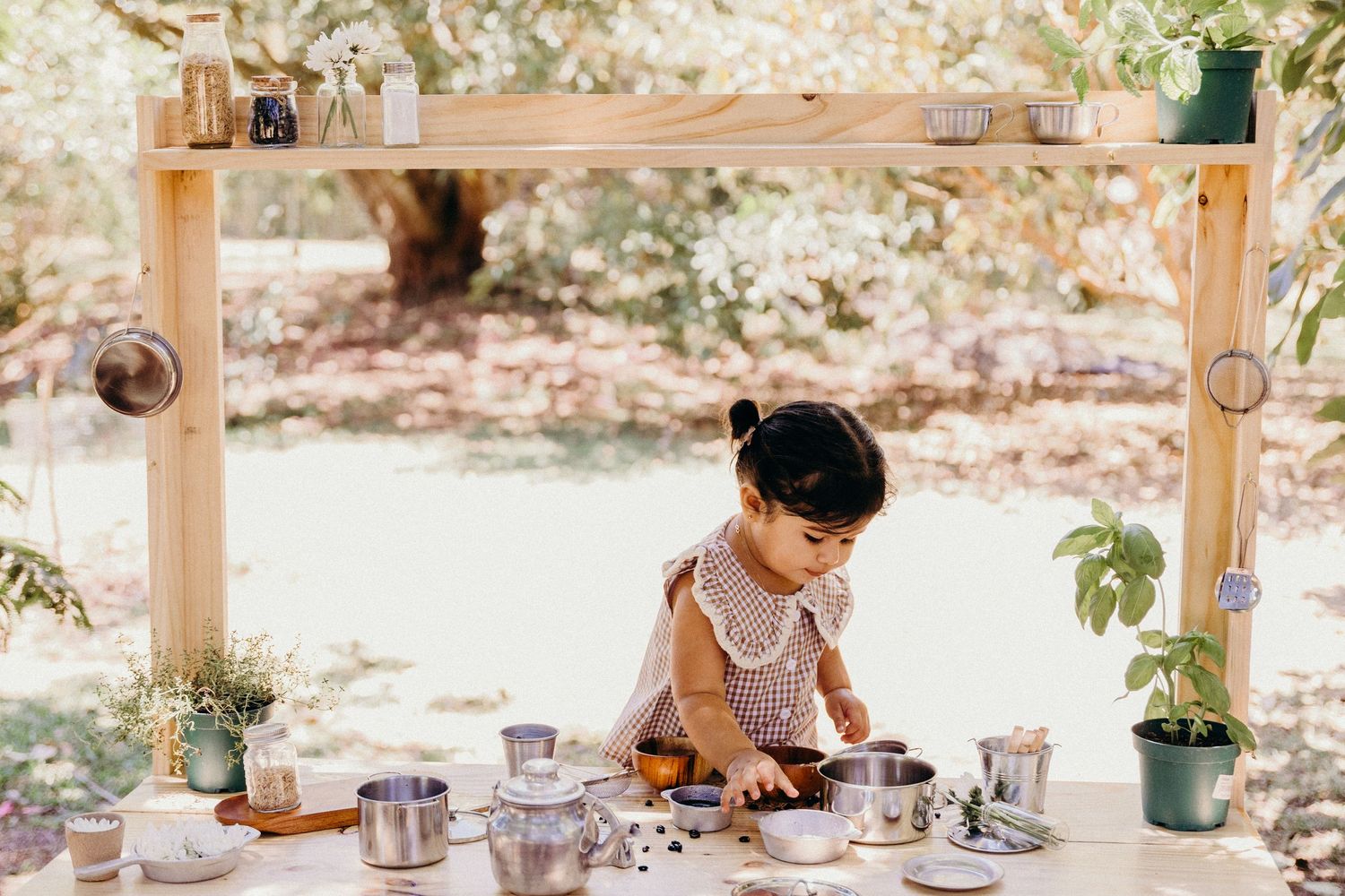 Toddler girl playing at an outdoor wooden mud kitchen with pots and pans surrounded by greenery.