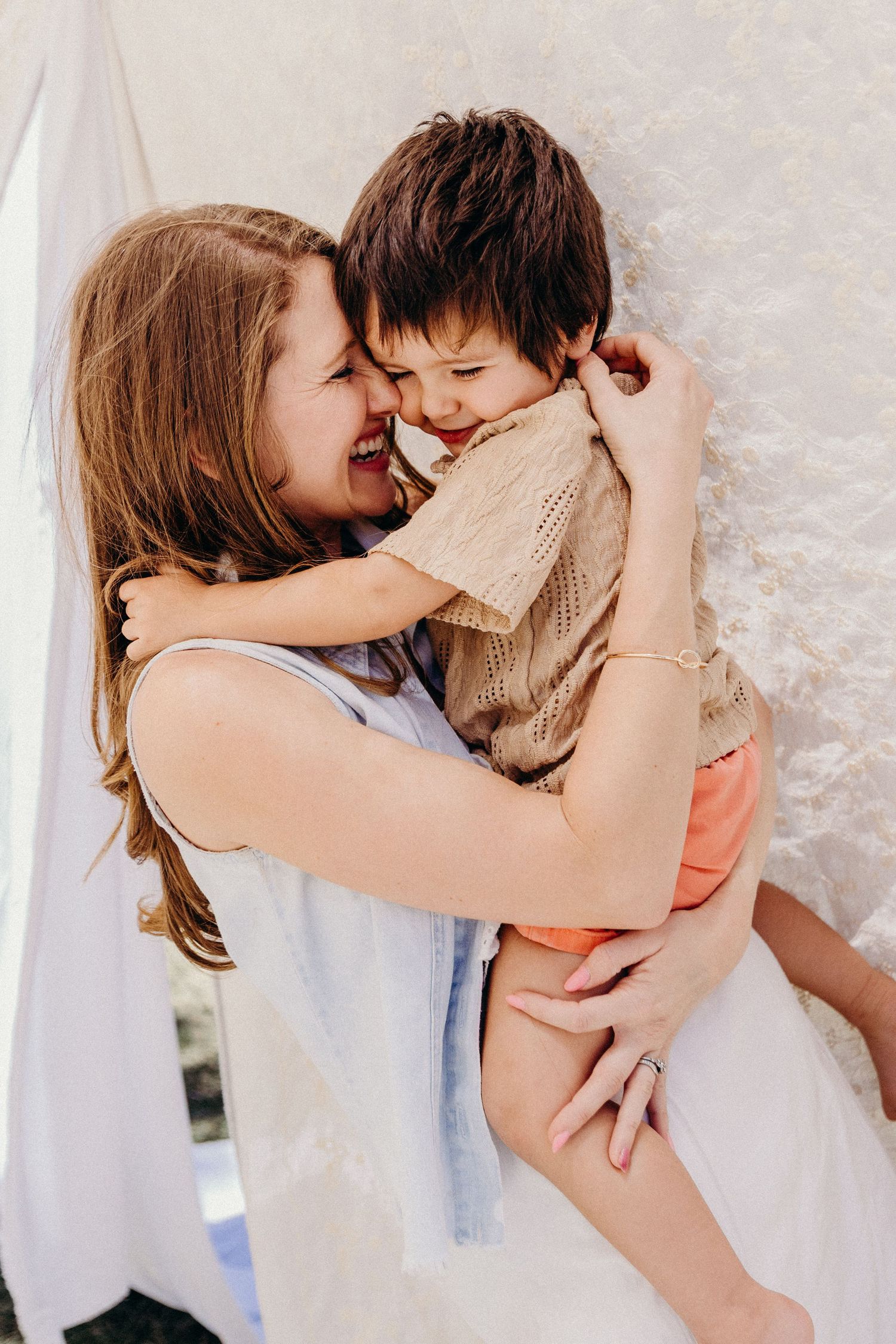 A joyful mother hugs and nuzzles her laughing young son outdoors, both sharing a tender, playful moment together.